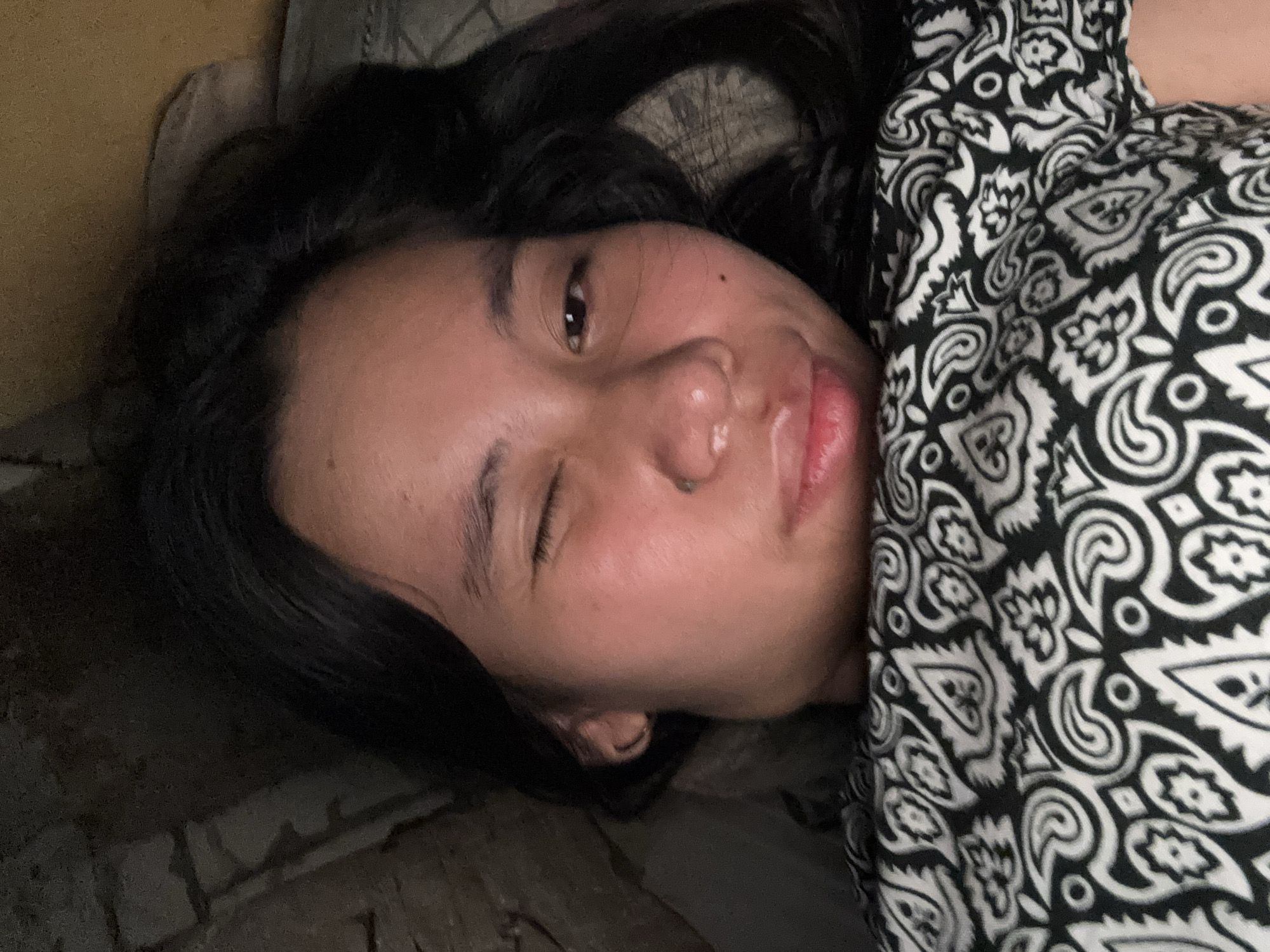 A young woman with dark hair is lying down, with her head partially visible. She is winking with her left eye, while her right eye is open. Her lips are slightly parted. She is wearing a black and white patterned garment, possibly a shirt or blanket. The background appears to be dark and possibly indoors, with some textured surfaces visible. The image does not provide enough information to determine the location in East Jakarta, Indonesia.
