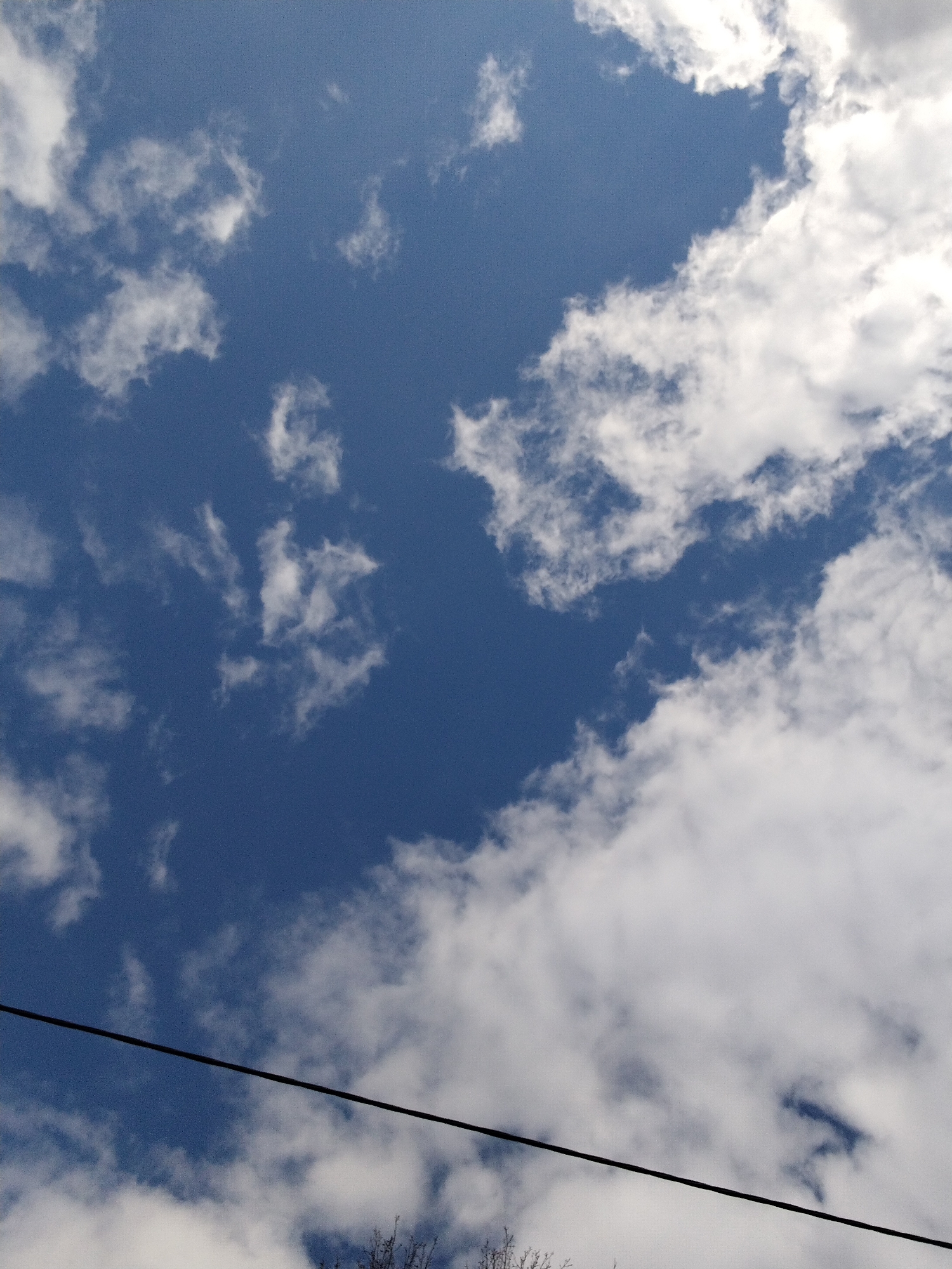 The image captures an upward view of a partially cloudy blue sky, indicative of daytime conditions. The sky is a vibrant, deep blue, interspersed with numerous white, fluffy cumulus clouds. Larger, denser cloud formations are visible on the right side and across the bottom of the frame, while smaller, more fragmented clouds are scattered throughout the top left and central areas. A thin, dark utility wire or cable runs horizontally across the lower section of the image. At the very bottom, the faint outlines of bare tree branches are visible, suggesting a season devoid of foliage, such as late winter or early spring. There are no people, specific activities, or text visible within the frame. The location context for this scene is Yaroslavl, Russia.