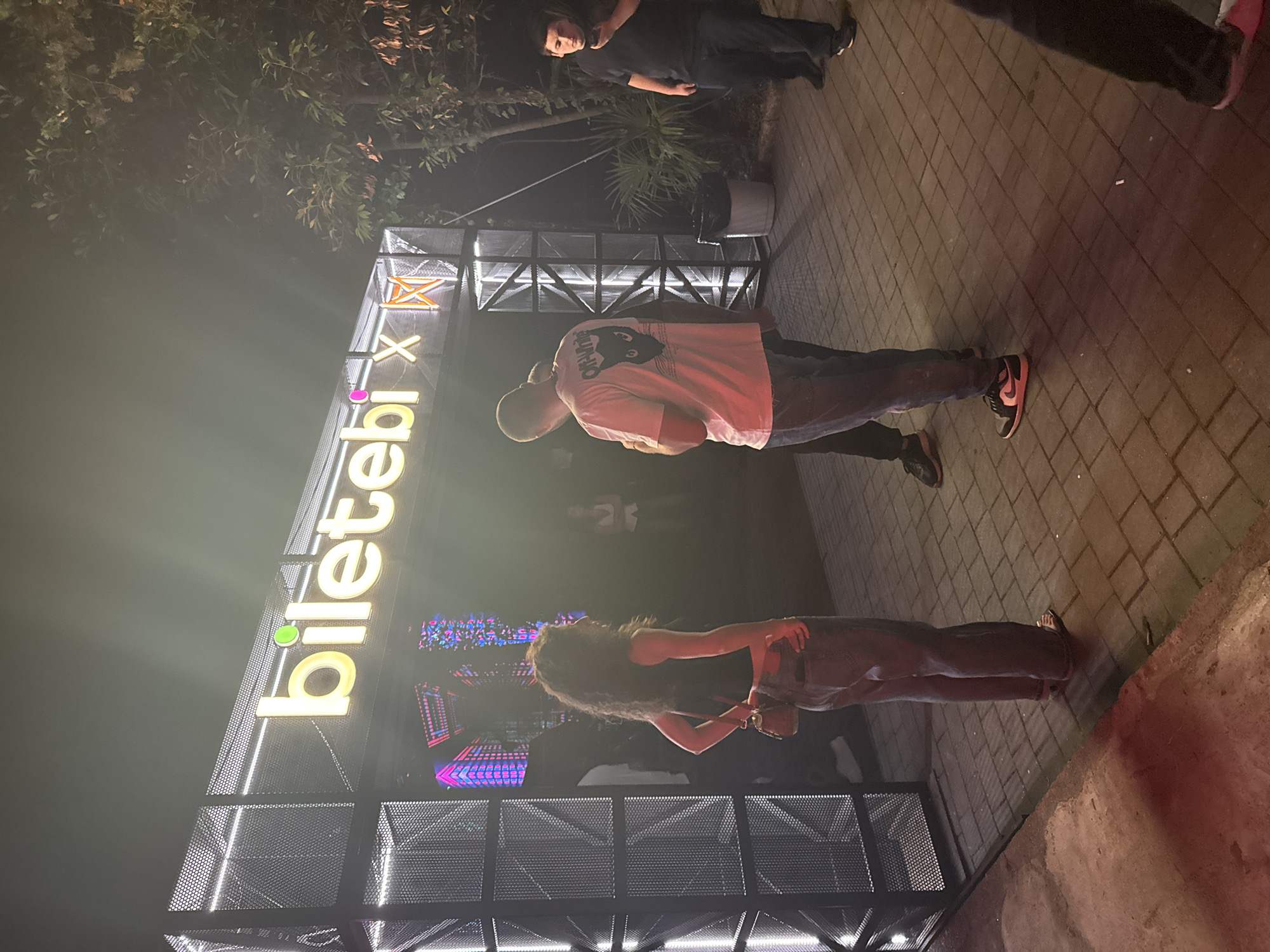 The image captures an outdoor scene at night, likely a nighttime event or venue entrance, featuring a large illuminated sign that reads "biletəbi X" in yellow neon letters with a green circular dot above the "t". The sign is set against a black metal frame structure adorned with white lights. In the foreground, three individuals are visible. A woman with curly dark hair, wearing a black sleeveless top and blue jeans, stands facing the right, holding a small brown purse. To her right, a man with a shaved head and a pink t-shirt with a black cat graphic on the back, also wearing blue jeans, stands with his back to the camera. Further to the right, a man in a dark t-shirt and jeans is looking at his phone. The ground is paved with interlocking bricks. Behind the main sign structure, a dark tree with some foliage is visible on the right side of the image. The overall lighting suggests an evening event in Batumi, Georgia.