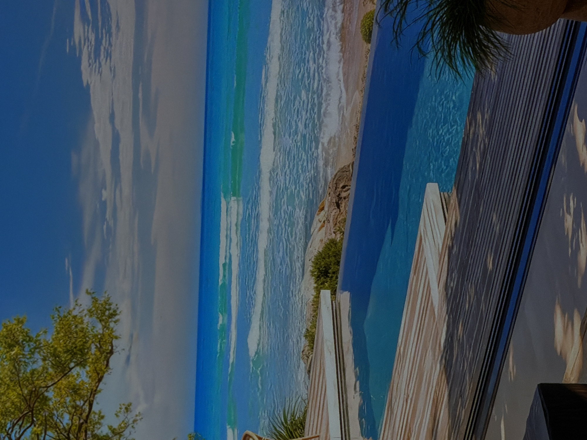 This media file presents a vibrant and picturesque coastal scene, likely a large mural or display panel, photographed in an interior setting in Sofiivska Borshchahivka, Ukraine. The image itself depicts a luxurious outdoor environment featuring an infinity pool overlooking a beach and the ocean under a bright, clear sky.
The foreground and middle ground on the right side are dominated by a dark-colored infinity pool with sparkling blue water. Adjacent to the pool is a light-colored wooden deck or pathway made of slats, creating a sense of a high-end resort. Green foliage, including palm fronds from a pot in the top right, and bushes near the pool's edge, add to the tropical feel.
Beyond the pool, a sandy beach is visible, with white-capped waves crashing gently on the shore. The ocean stretches out to the horizon, showcasing various shades of blue and turquoise, indicating clear waters. The coastline features some rocky formations interspersed with green vegetation.
The upper-left portion of the image displays a bright blue sky dotted with scattered white clouds, suggesting a sunny day with pleasant weather. In the bottom-left corner, the vibrant green canopy of a tree adds natural elements to the composition.
No people are visible in the scene, and no specific activity is taking place. The overall mood is one of serenity and an idyllic vacation escape. There is no visible text within the image.