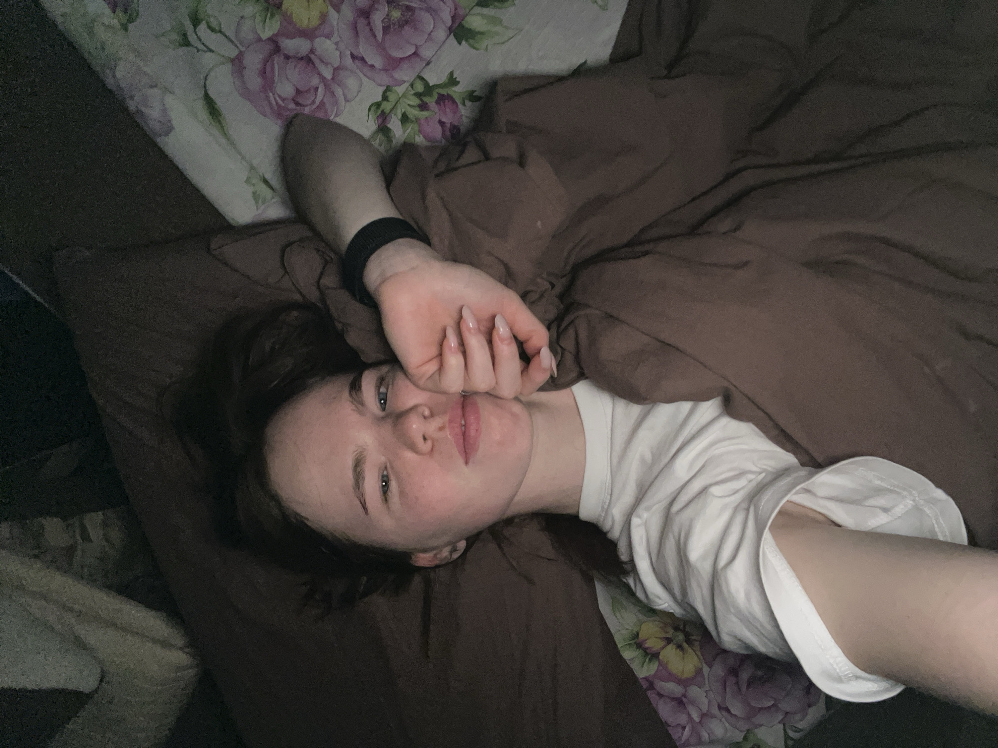 Location: An indoor residential setting in Chaikovsky, Russia.
Scene: A young adult female is depicted lying on her back in a bed, looking directly at the camera. She possesses fair skin, blue eyes, and dark hair. She is dressed in a white short-sleeved t-shirt. A black elastic wristband is worn on her left wrist. Her fingernails are long, manicured, and coated with a light-colored polish. She is partially covered by a dark brown duvet or blanket, which appears wrinkled. Her head rests on a dark brown pillow. To her right, a segment of a white pillowcase or sheet featuring a floral pattern of pink and purple roses with green leaves and yellow elements is visible. The overall lighting within the scene is dim.