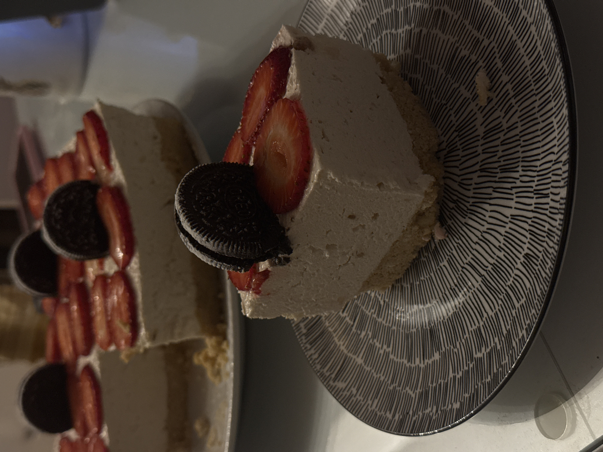 A slice of cake sits on a plate with a black and white pattern of concentric lines. The cake is topped with sliced strawberries and an Oreo cookie. Another slice of cake is on a plate to the left. The background is out of focus. This could be a dessert served at a cafe or restaurant in Bar, Montenegro, but the location cannot be confirmed without additional information.