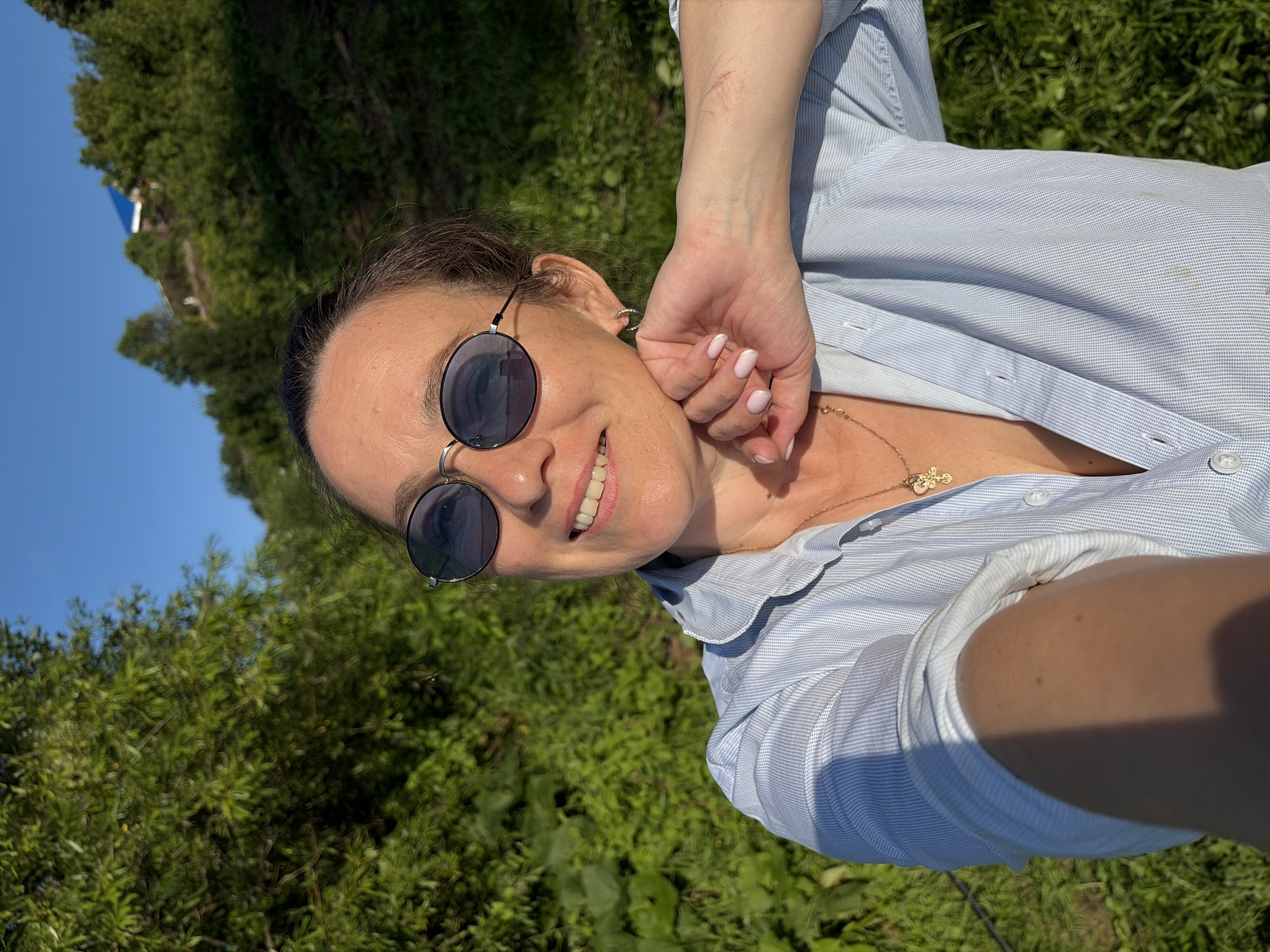 A woman is taking a selfie in a rural area. She is wearing sunglasses and a light blue, pinstriped button-down shirt. She has her hand on her chin and is smiling at the camera. The background is filled with lush green foliage and a bright blue sky. A small, blue-roofed building is visible on a hill in the distance. The scene appears to be located in Olkhovka, Russia, given the natural landscape.
