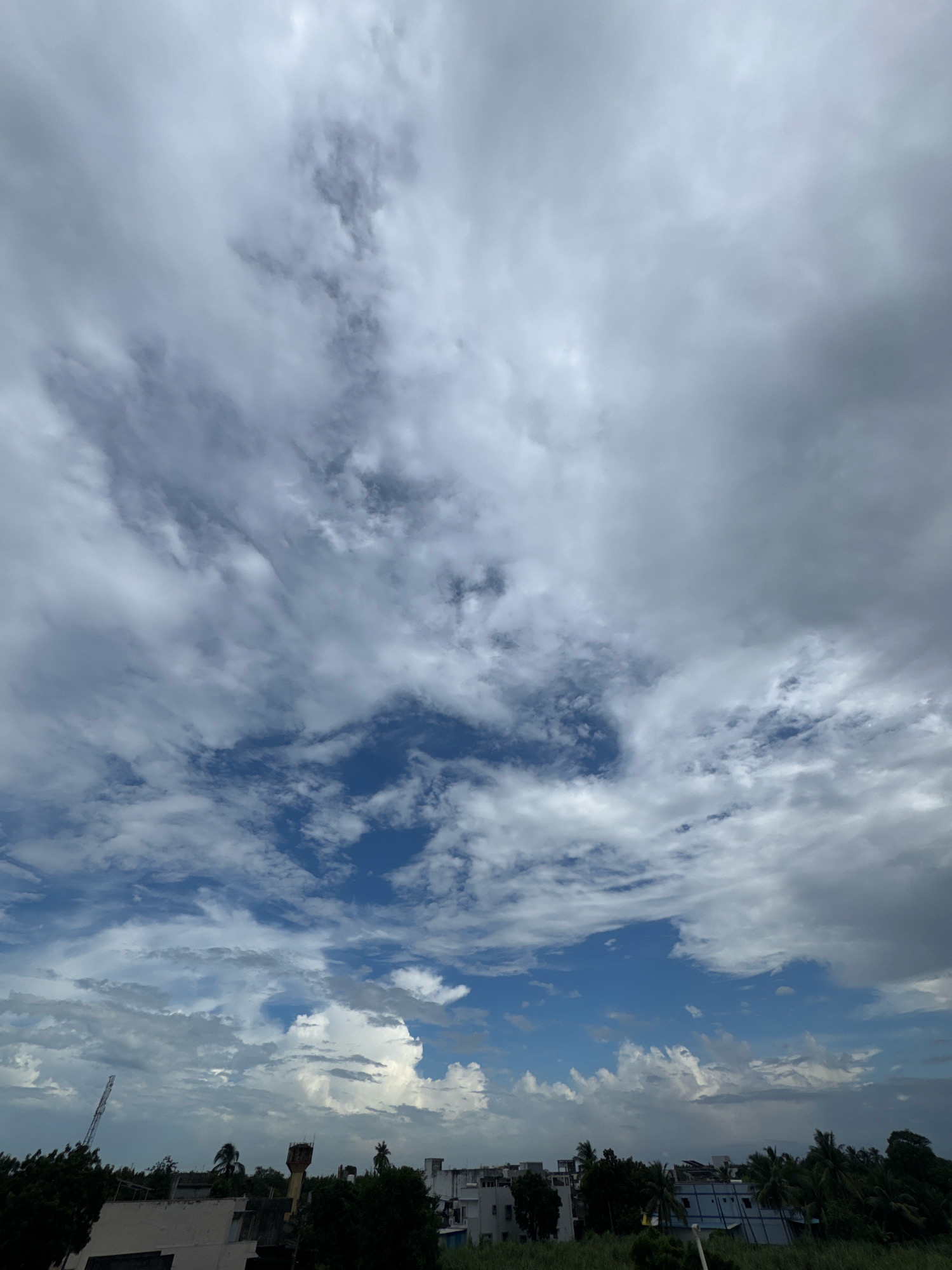 The image shows a sky filled with a mix of white and gray clouds, with patches of blue sky visible. In the distance, there is a low-rise urban landscape, likely in the city of Jokha, India. The horizon is lined with buildings, trees, and a tall structure that appears to be a communication tower or crane. The overall scene suggests a cloudy or overcast day, possibly before or after rain.