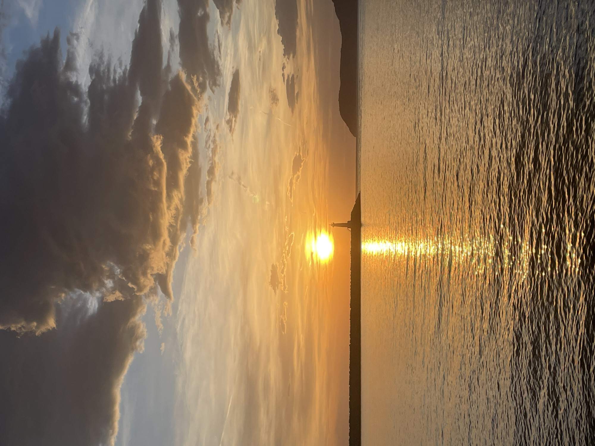 A scenic view features a sunlit body of water that extends horizontally across the scene. Reflections from the sun create a sparkling effect on the water's surface. In the background, a land formation appears to be situated either within the water or along the coast. Above, a dramatic sky with layered clouds adds depth to the composition. The sunset over the sea is captured at Bar in Montenegro.