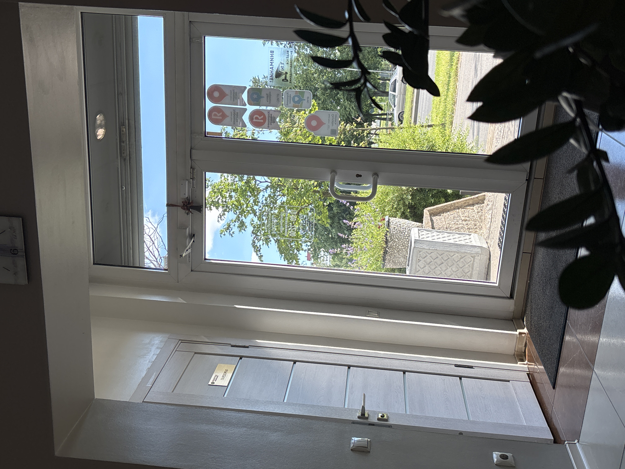 This is a medium shot, taken from inside a building, looking through a glass door to the outside. The interior is dimly lit, with a gray wooden door in the foreground. The door has a small sign on it that reads "ГАРДЕРОБ" (wardrobe/cloakroom). There is a metallic handle and lock mechanism on the door.
The glass door leads outside to a bright, sunny day. Green trees and bushes are visible, with a paved area and a road in the background. A light-colored car is parked on the roadside. The sunlight creates strong reflections on the glass, obscuring some details of the exterior.
The glass door itself has several stickers or decals on it. Some are circular with a red pin icon and text in a foreign language, possibly Cyrillic. Other stickers are rectangular, with text and icons. One sticker is predominantly white with blue and red elements.
The scene appears to be an entrance to a building, possibly an office or a public establishment. The lighting suggests it is daytime, likely late morning or early afternoon given the brightness of the sun. No people are visible. The overall impression is of a transitional space between indoors and outdoors on a clear day.