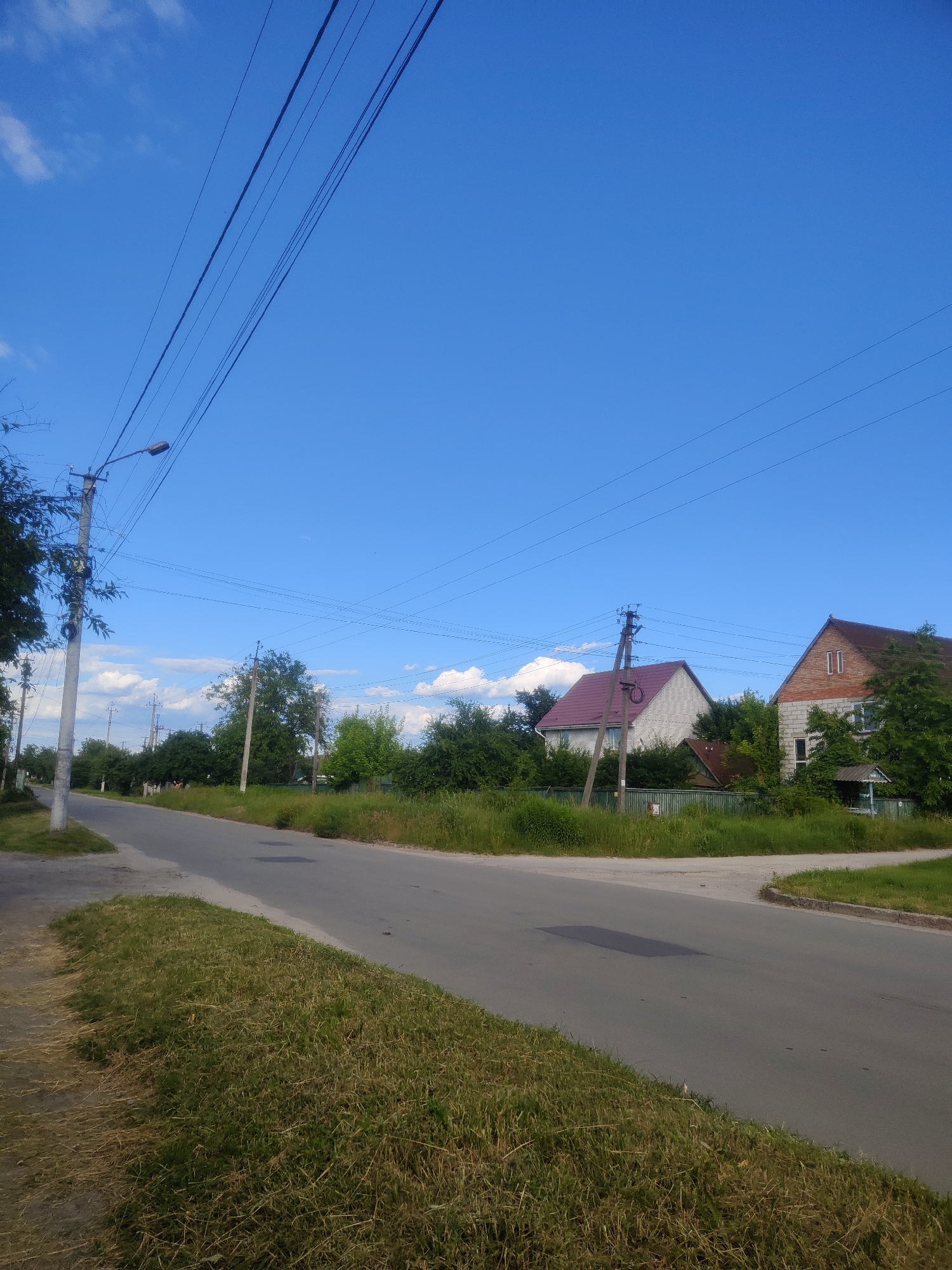 The image shows a street scene in Borova, Ukraine. A paved road curves to the right, bordered by green grass on both sides. Several houses with red roofs are visible behind a green fence. Power lines crisscross the bright blue sky, supported by utility poles. The overall impression is a quiet, rural street on a clear day.
