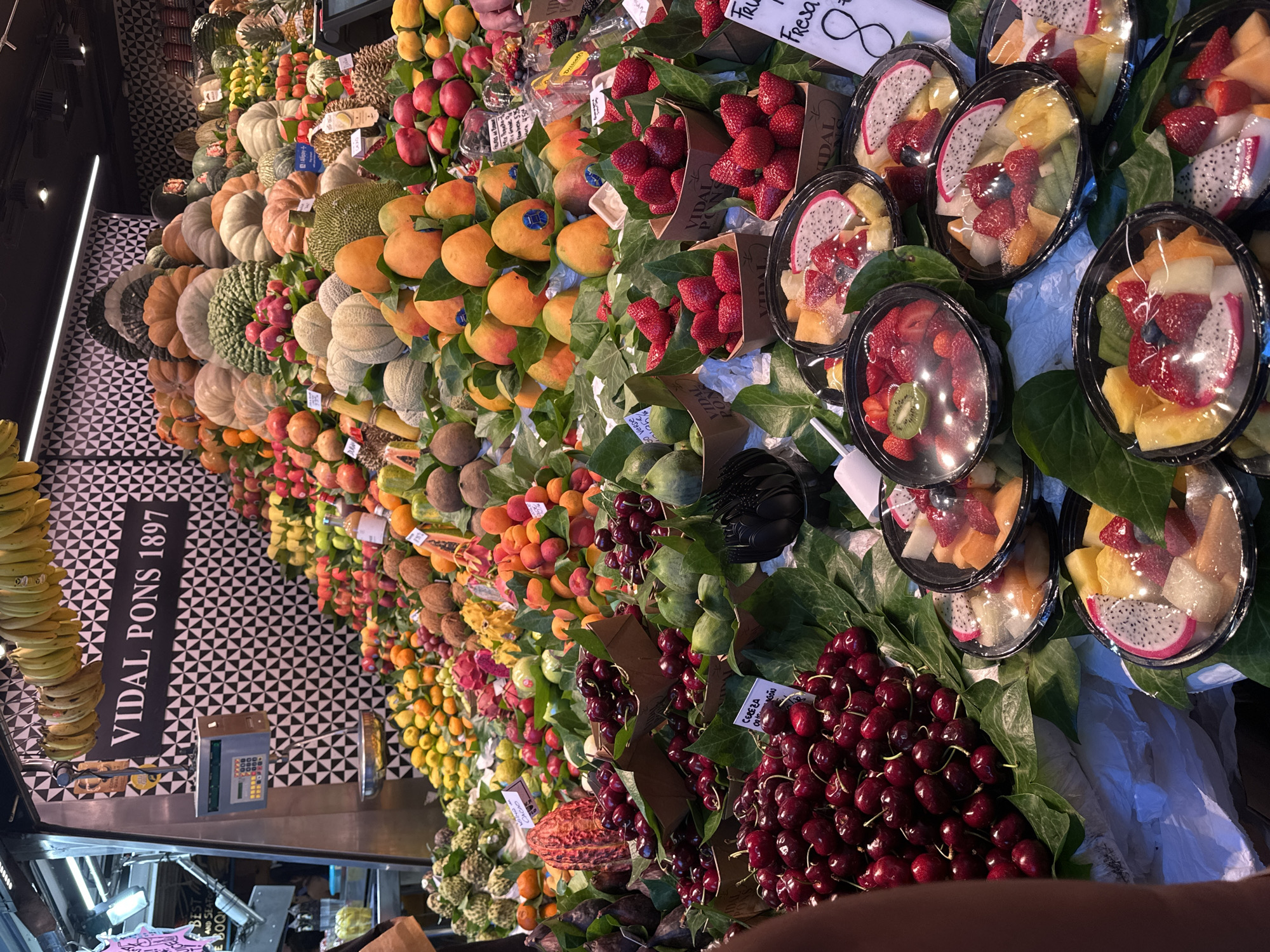 This image shows a vibrant fruit stall in La Boqueria Market, located on La Rambla in Barcelona, Spain. The stall is overflowing with a colorful assortment of fresh fruits, meticulously arranged on display. Prominently featured are large piles of dark red cherries in brown paper containers, surrounded by green leaves. To the right, various fruit salads are packaged in clear plastic containers, showcasing a mix of dragon fruit, strawberries, pineapple, and other tropical fruits. In the background, a large display of pumpkins and squashes is visible, along with bunches of bananas hanging overhead. The stall's signage, "VIDAL PONS 1897," suggests it is a long-standing establishment. The overall scene depicts a bustling marketplace, abundant with fresh produce and capturing the lively atmosphere of a Spanish food market.