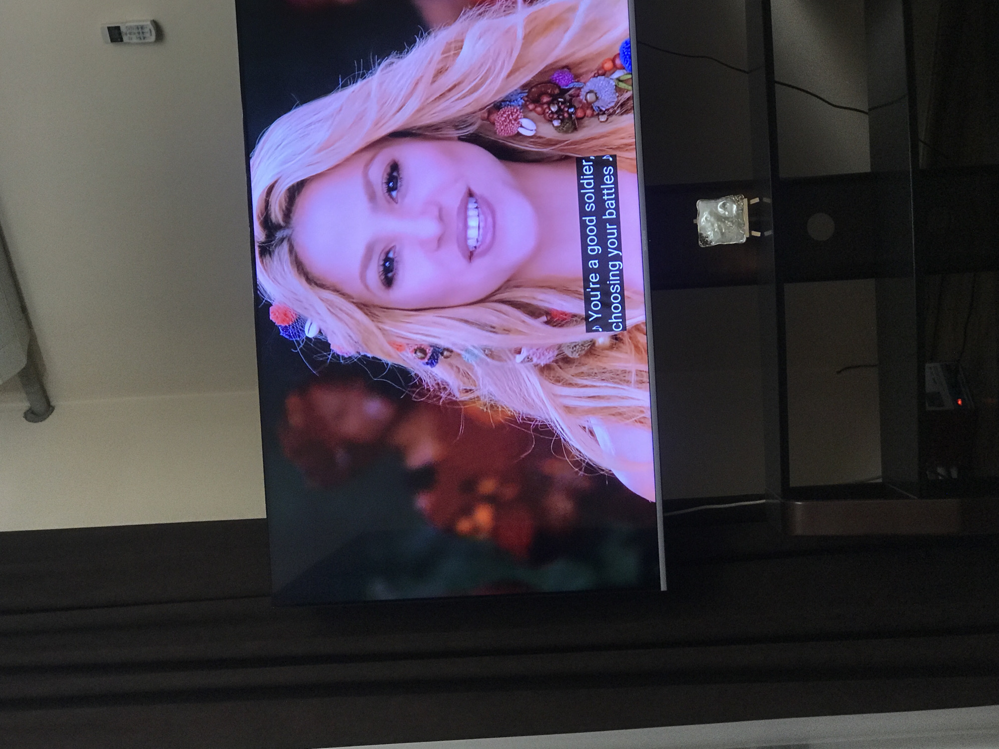 A television displaying a close-up of a woman with fair skin, blonde hair, and decorative accessories in her hair. The woman is smiling and the screen displays the text: "You're a good soldier, choosing your battles". The television is mounted on a black shelving unit. In the background, a wall can be seen along with a remote control. The setting is inside, likely a residential interior. There is no information available in the image to determine the location as Yerevan, Armenia.