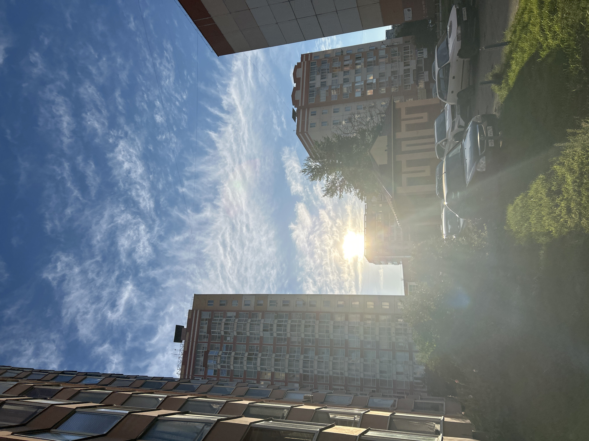 The image depicts a residential area in Tomsk, Russia, during daylight. The scene is dominated by tall apartment buildings and a clear blue sky with wispy white clouds. Several cars are parked in a lot in the foreground. The sun is visible and shining brightly, causing a lens flare in the lower portion of the image. The buildings are of a modern design, with visible windows and balconies. The overall impression is of a typical urban environment during a sunny day.