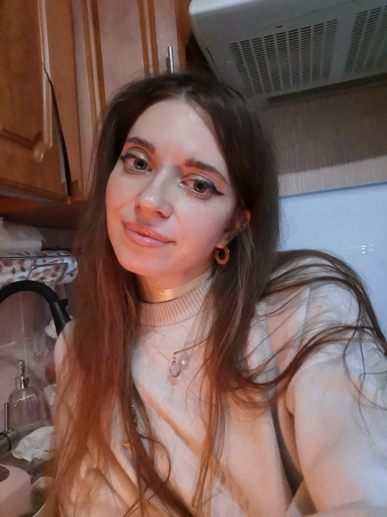 A young woman with long brown hair is posing for a selfie in what appears to be a kitchen. She has light skin, dark eyeliner, and glossy lips. She is wearing a cream-colored sweater, a gold choker, and gold hoop earrings. A necklace with a pendant hangs below the choker. Behind her are wooden cabinets, a microwave, and various kitchen items. The location is Unknown, Unknown.
