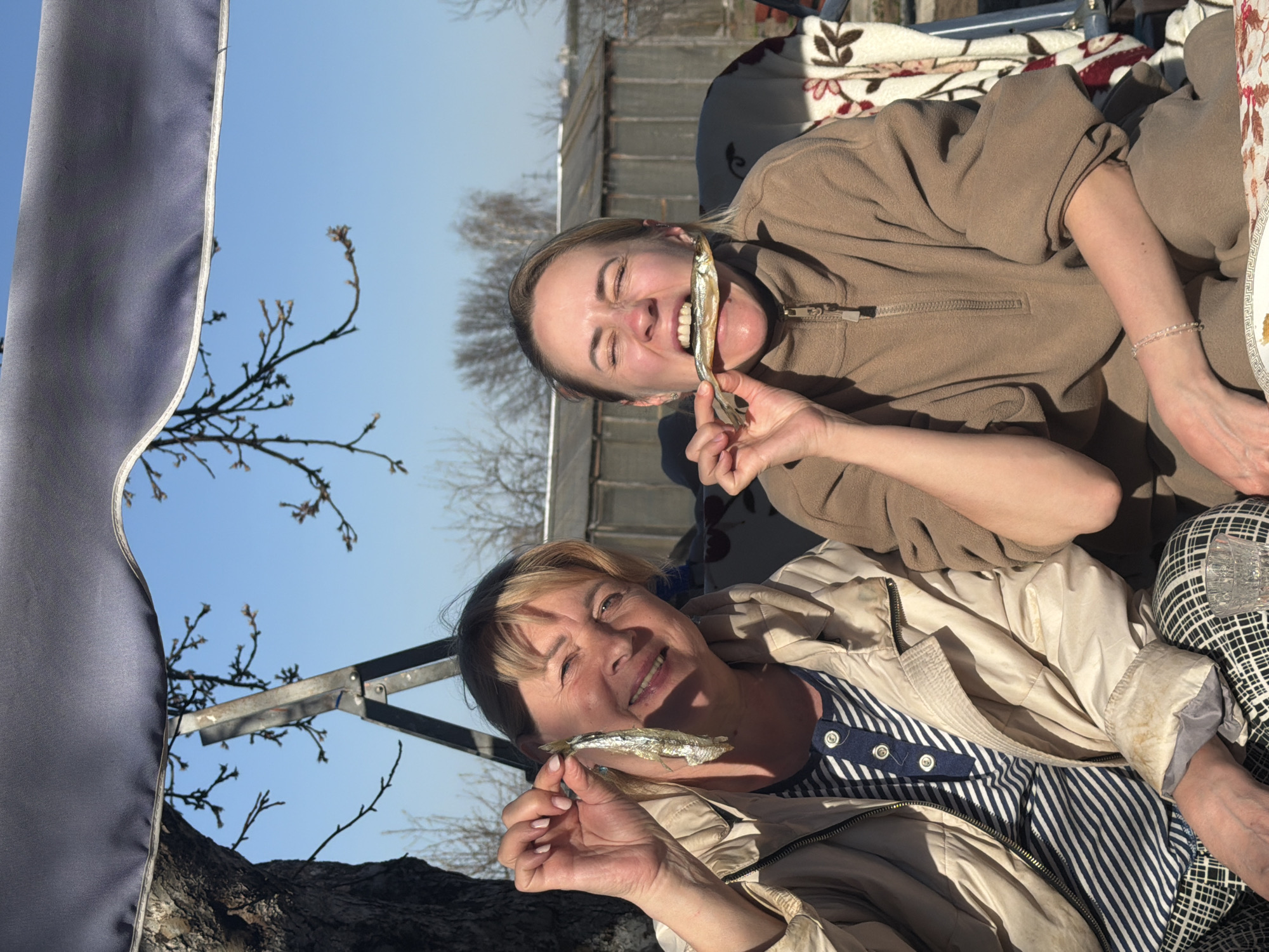 Two women are sitting outside, possibly on a swing or outdoor furniture, holding small, dried fish. The woman on top is younger, with her eyes closed and a fish in her mouth. The woman below has short hair and is smiling, holding a fish in her hand. They are dressed in casual clothing. The background includes trees, a building, and a blue sky. The location is Chaikovsky, Russia.