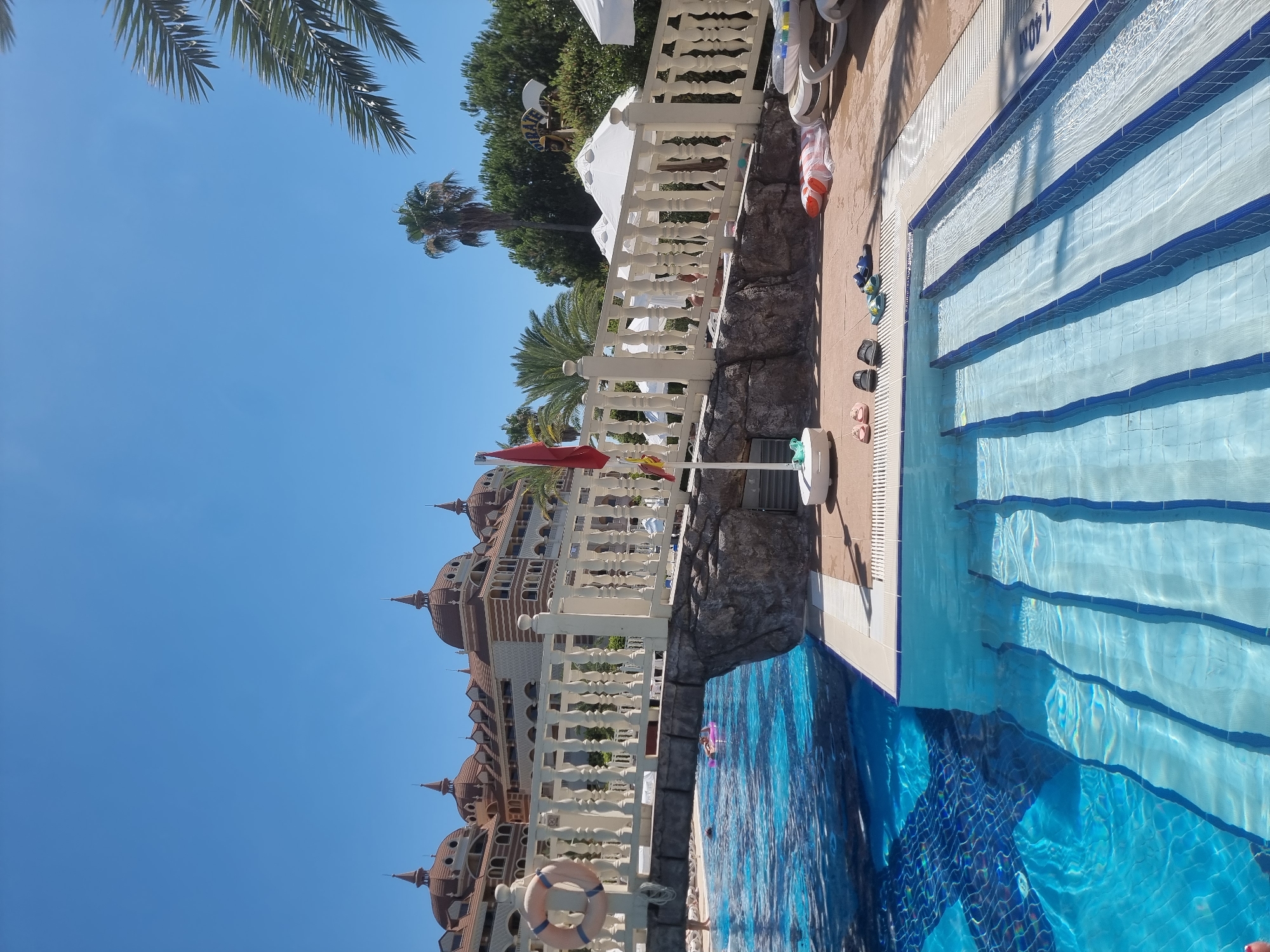 The image captures a sunny day at a resort in Manavgat, Turkey. The scene is dominated by a large, clear blue swimming pool with multiple steps leading into it. The water is sparkling, indicating bright sunlight. Adjacent to the pool is a paved area with several pairs of flip-flops and sandals scattered around, suggesting a relaxed, vacation atmosphere. A white railing with ornate balusters runs along the edge of the pool area, separating it from a rocky embankment.
In the background, a grand building with distinctive dome-shaped roofs and intricate architectural details stands tall against a clear, vibrant blue sky. Palm trees are visible, with one particularly tall one casting a shadow. A red umbrella is partially visible near the railing, and a life preserver ring is attached to the railing in the foreground.
In the pool, a person can be seen swimming with a pink inflatable float. The overall mood is one of leisure and enjoyment, typical of a holiday resort on a warm, sunny day. The time of day is clearly midday, given the strong sunlight and shadows. The location cues, such as the architecture and palm trees, combined with the provided context, confirm the setting as Manavgat, Turkey. There is no visible text in the image.