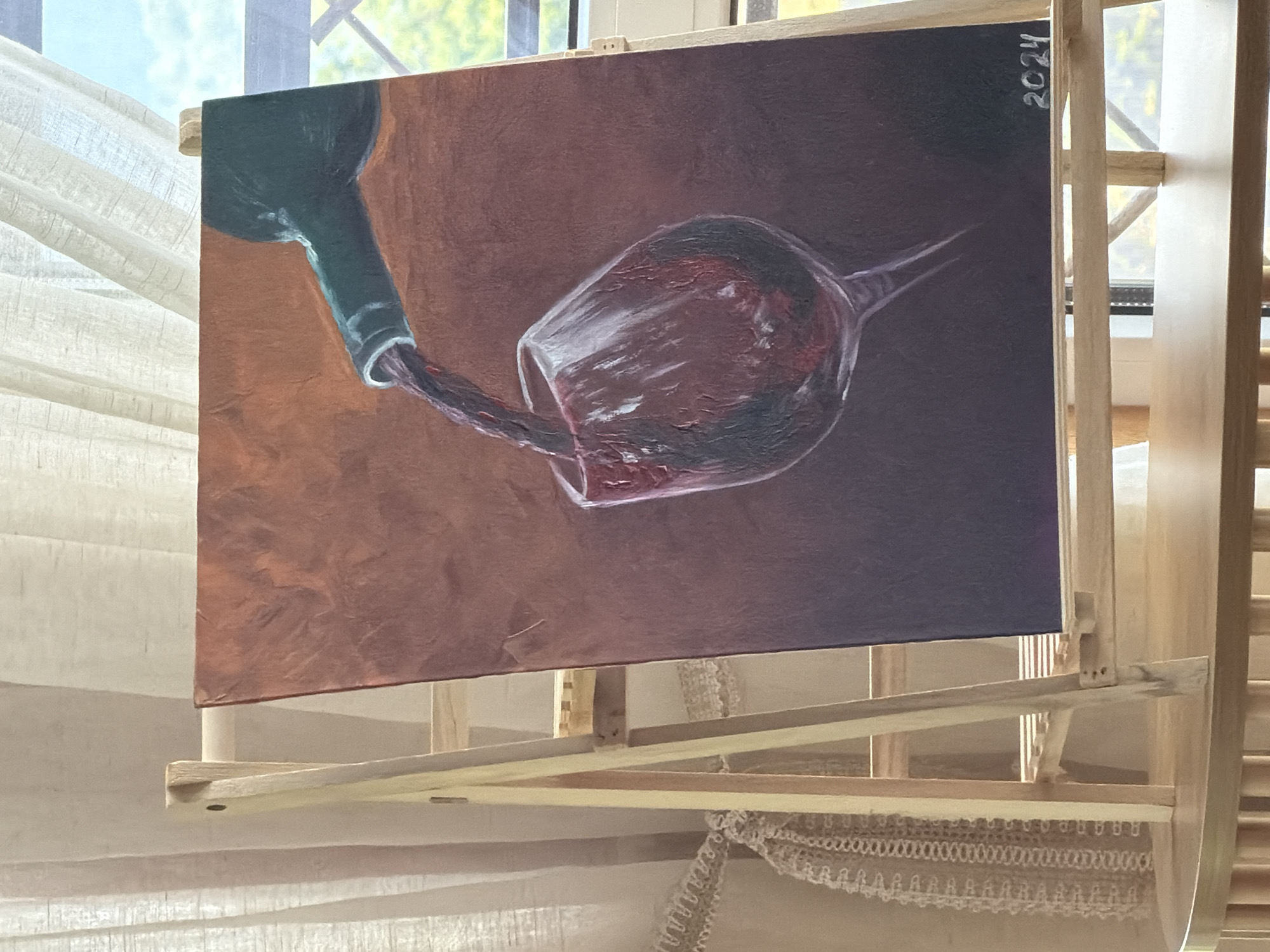 The image shows an oil painting of red wine being poured from a dark green bottle into a glass, displayed on a wooden easel. The painting has a textured appearance and features warm, deep tones of red, brown, and dark purple. The bottle's neck and the stream of wine are prominently rendered, with highlights suggesting the sheen of the liquid. The glass is partially filled with wine, reflecting light and showcasing the rich color. In the bottom right corner of the painting, the year "2024" is inscribed in white. The easel is positioned in front of a window with sheer curtains, through which a blurry outdoor scene with greenery is visible. The overall setting appears to be indoors, possibly a home or studio, bathed in natural light. No people are visible in the image, and there is no information or visual evidence to suggest the location is the city of Nizhnyaya Shilovka, Russia.