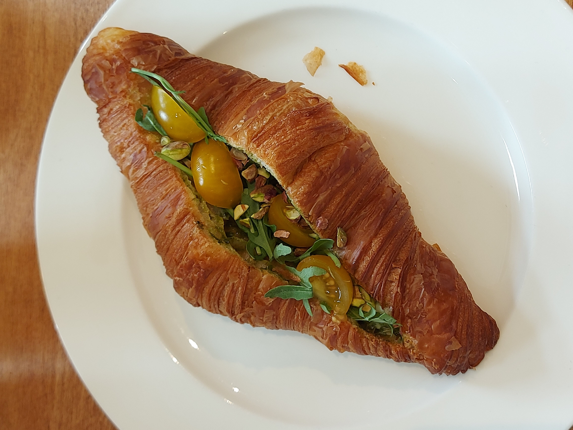 A croissant sandwich with yellow tomatoes, greens, and nuts on a white plate is photographed from above. The flaky, golden-brown croissant is split open lengthwise, revealing a filling of green pesto-like spread. Cut yellow tomatoes, small green leaves, and chopped nuts are visible in the filling. Two small, light-brown crumbs sit to the right of the sandwich on the plate. The plate rests on a wooden surface. No people are visible. The location is unknown; there is no indication of Saint Petersburg, Russia.