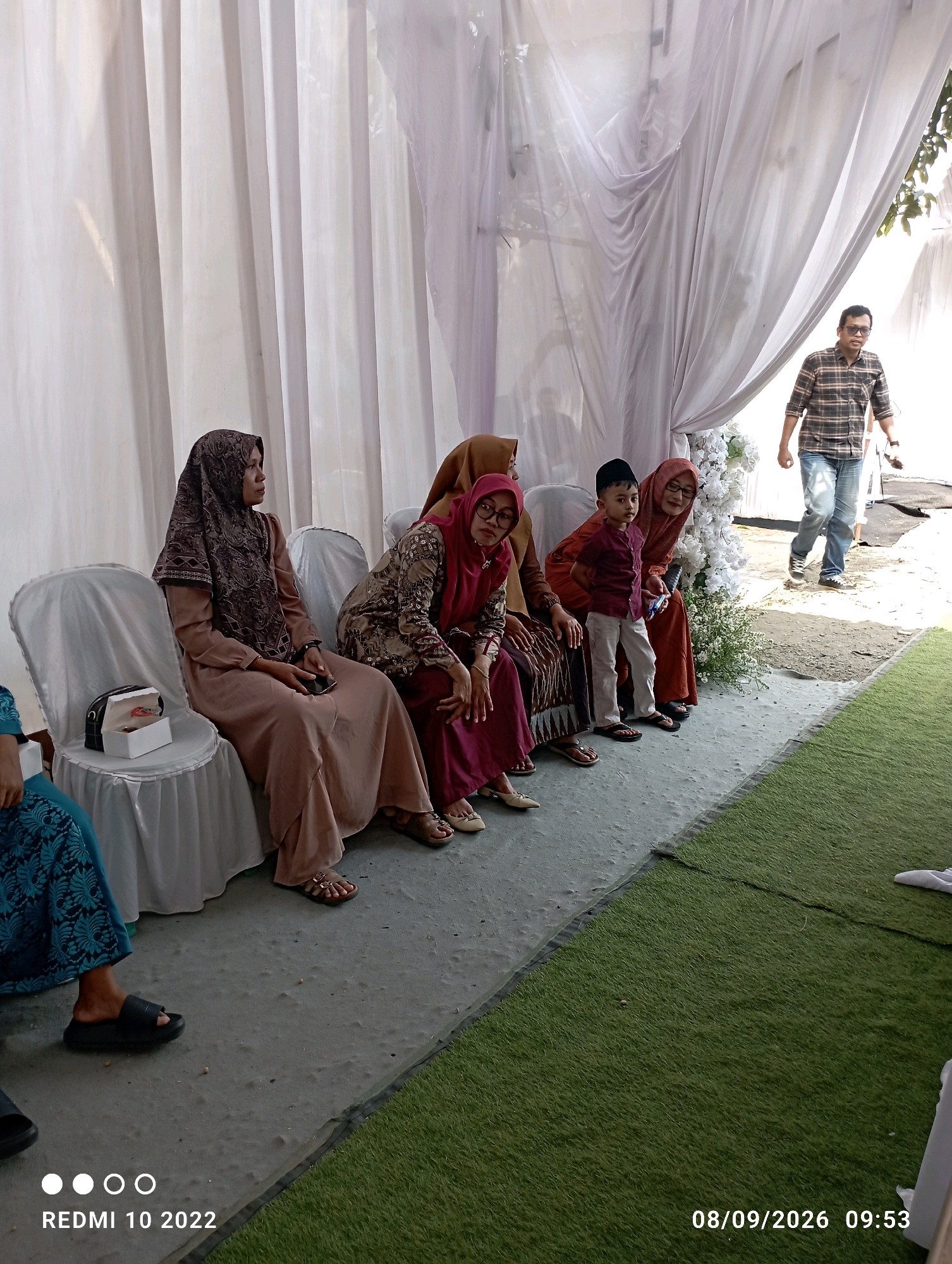 In Dukuhseti, Indonesia, a gathering takes place under a white canopy. Four women are seated on white chairs, facing left, their heads adorned with hijabs. The woman on the far left wears a brown hijab and a matching long-sleeved dress. To her right, another woman in a patterned dress and a maroon hijab looks towards the camera. The third woman, also in a maroon hijab, is seated next to a young boy. This boy, wearing a maroon shirt and white pants, stands slightly apart, facing forward. The fourth woman, wearing an orange hijab, is seated beside the boy. Further right, a man in a plaid shirt and jeans walks away from the group. The ground is a mix of grey concrete and artificial green turf. A white box with gold trim sits on the chair to the left of the seated women. The scene appears to be an outdoor event, possibly a reception or a social gathering. The timestamp "08/09/2026 09:53" is visible in the lower right corner.