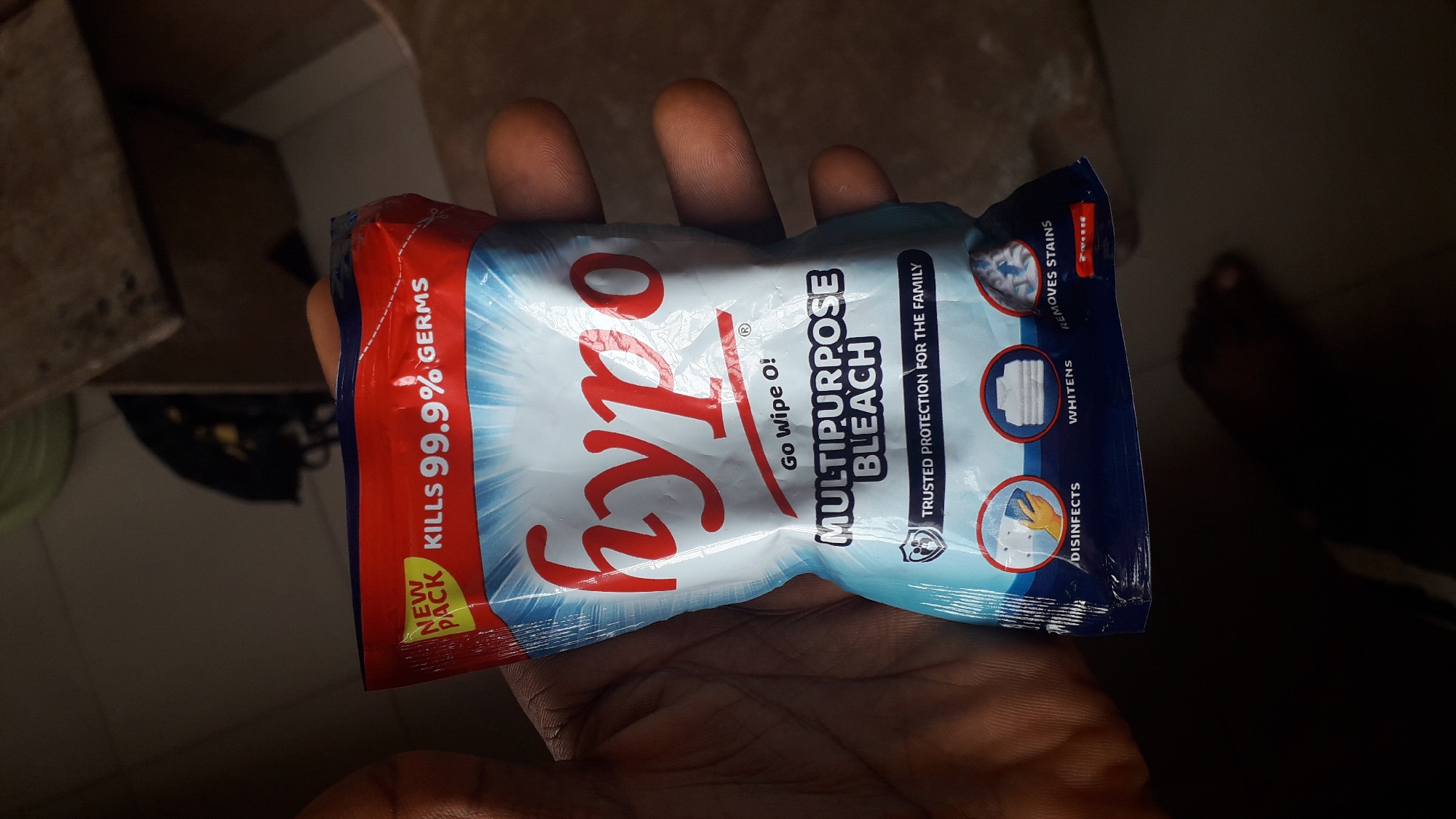 A close-up shot features a hand holding a small, sealed packet of "Hypo Multipurpose Bleach," with the packaging predominantly red, white, and blue. The packet's details include "Kills 99.9% Germs," "Go Wipe O!," "Trusted Protection for the Family," and icons indicating disinfection, whitening, and stain removal. The hand holding the product appears to be resting in an indoor environment, but specific details of the location in Nkpor, Nigeria are not discernable from the image.