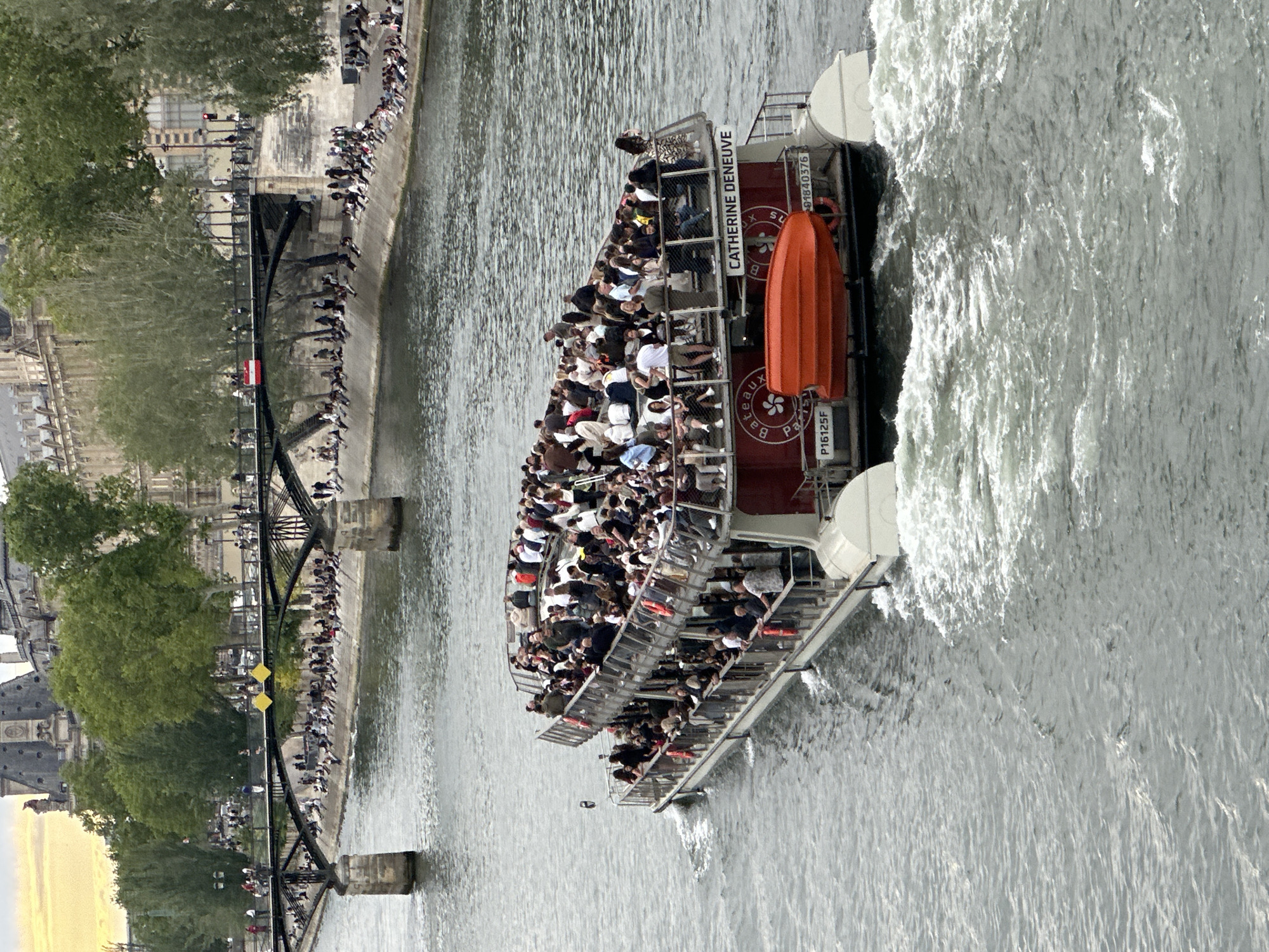 The image depicts a sightseeing boat, filled with passengers, navigating the Seine River in Paris, France. The boat, named "Catherine Deneuve," creates a noticeable wake as it moves through the water. A pedestrian bridge spans the river, with numerous people lining its sides. The architecture of Paris is visible along the riverbanks, including historical buildings. The scene suggests a typical day of tourism and leisure activities in the city. The overall environment is urban and watery.