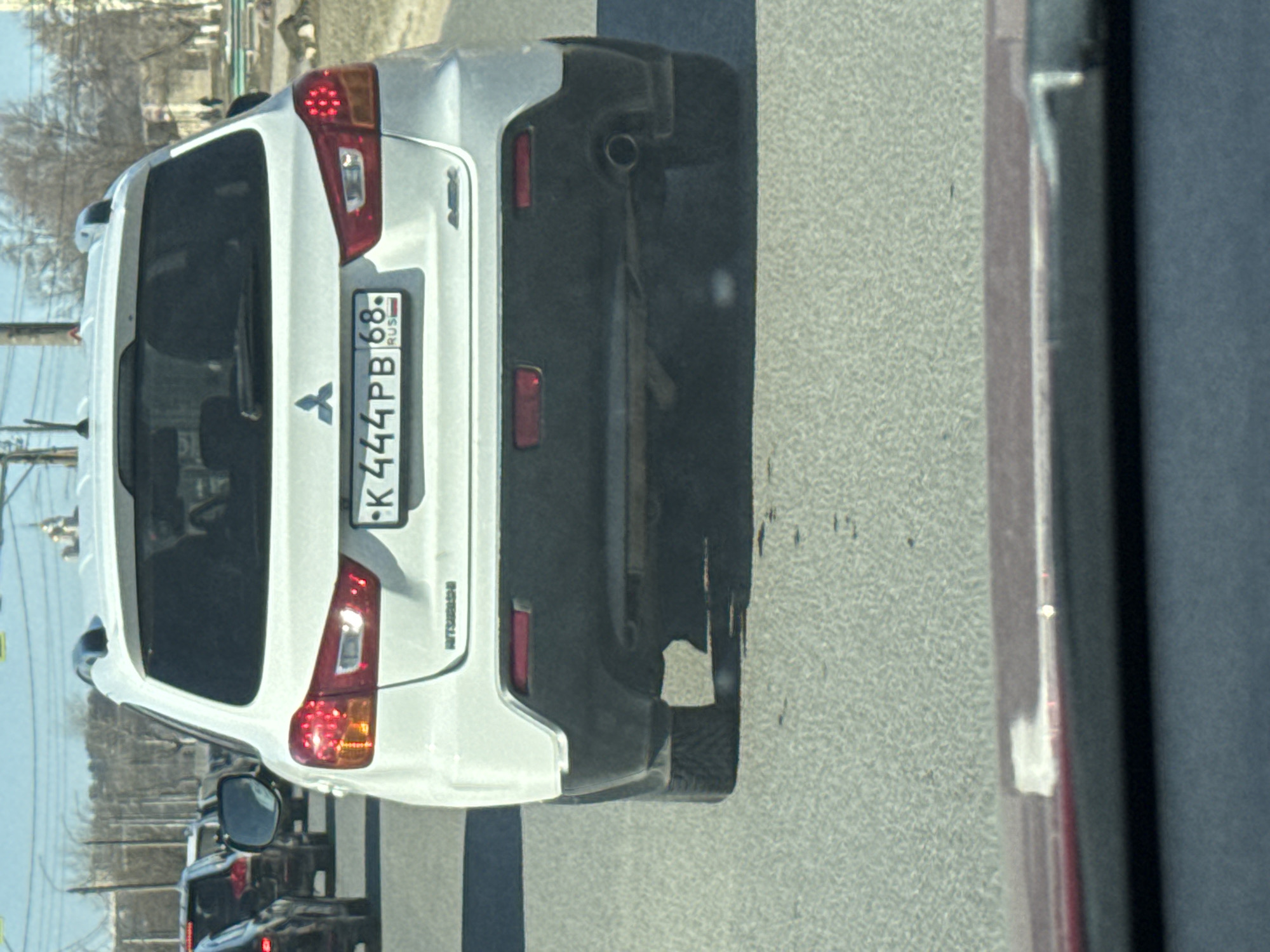 The image shows the rear of a white Mitsubishi car with the Russian license plate K444PB 68. The number 68 indicates the Tambov Oblast region of Russia. The car is on a road, likely in a city based on the surrounding environment. Other cars are visible behind it. The image appears to have been taken from another car. Based on the details available, it is not possible to verify the location in the city of Vladimir.