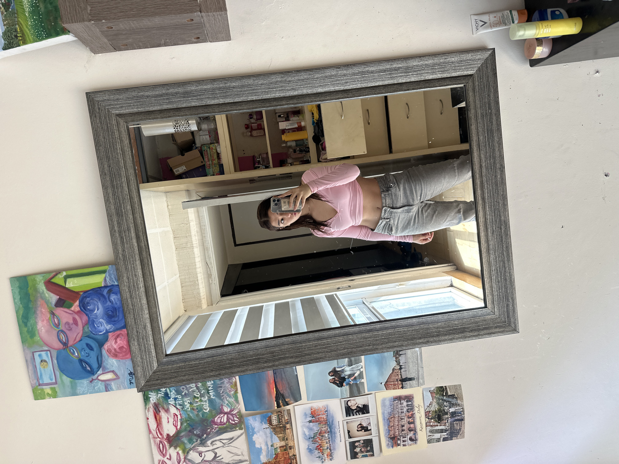 A young woman is taking a selfie in a mirror. The mirror is hanging on a wall decorated with artwork and postcards. The reflection in the mirror shows a room with a dresser and a window. Some toiletries are placed on the wall, above the mirror. The postcards feature scenes of cities such as Warsaw and Kazimierz Dolny.