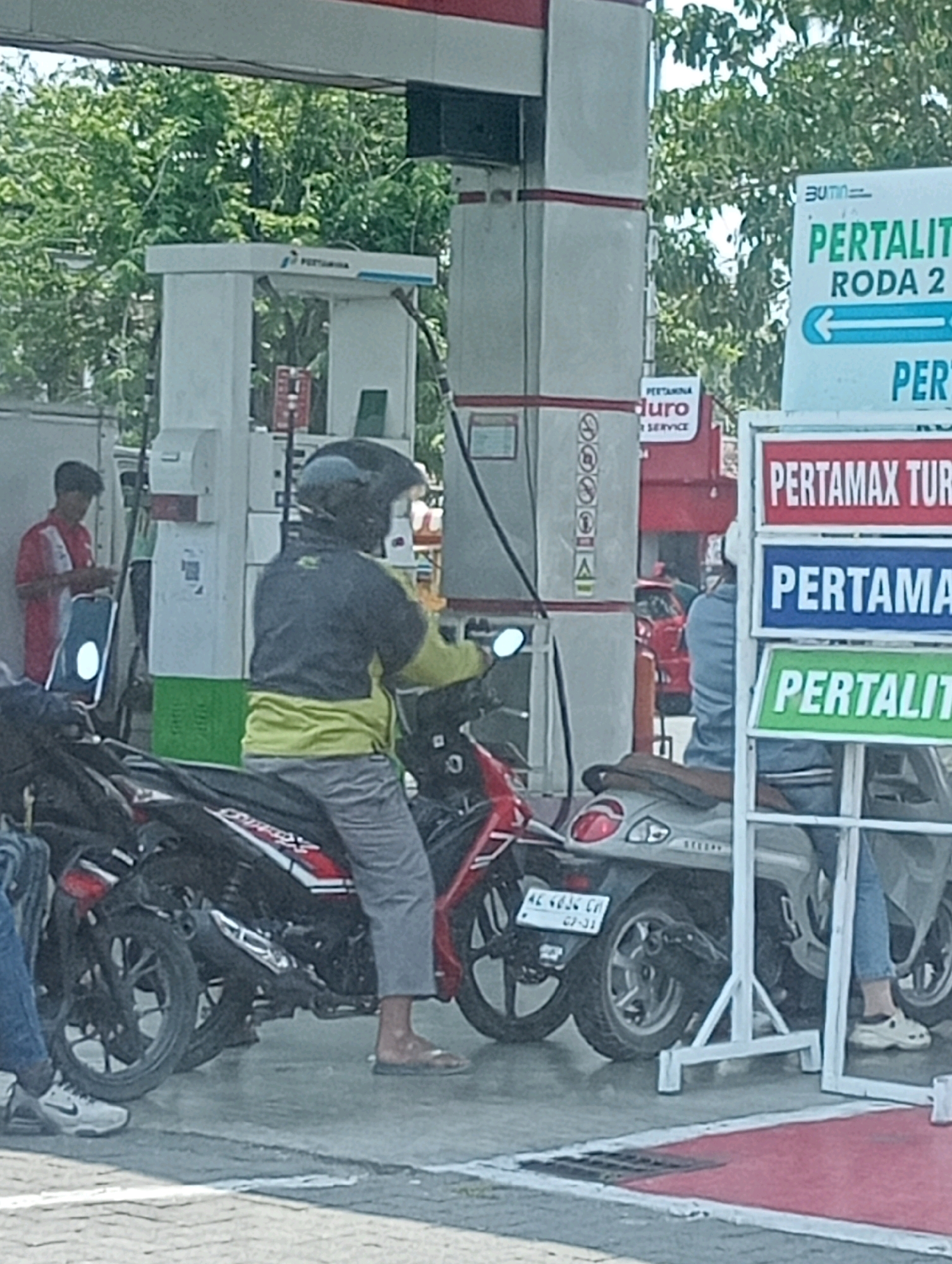At a Pertamina gas station in Plosojenar, Indonesia, a person wearing a black helmet and a yellow and grey jacket is seated on a motorcycle, waiting for service. Another person in a red shirt stands near a gas pump, holding a tablet. The scene is bustling with activity, with several other motorcycles and people present, suggesting a busy day at the station. The gas station is equipped with multiple pumps and signage displaying fuel prices and types. The environment appears to be an outdoor setting, possibly a street or roadside location.
