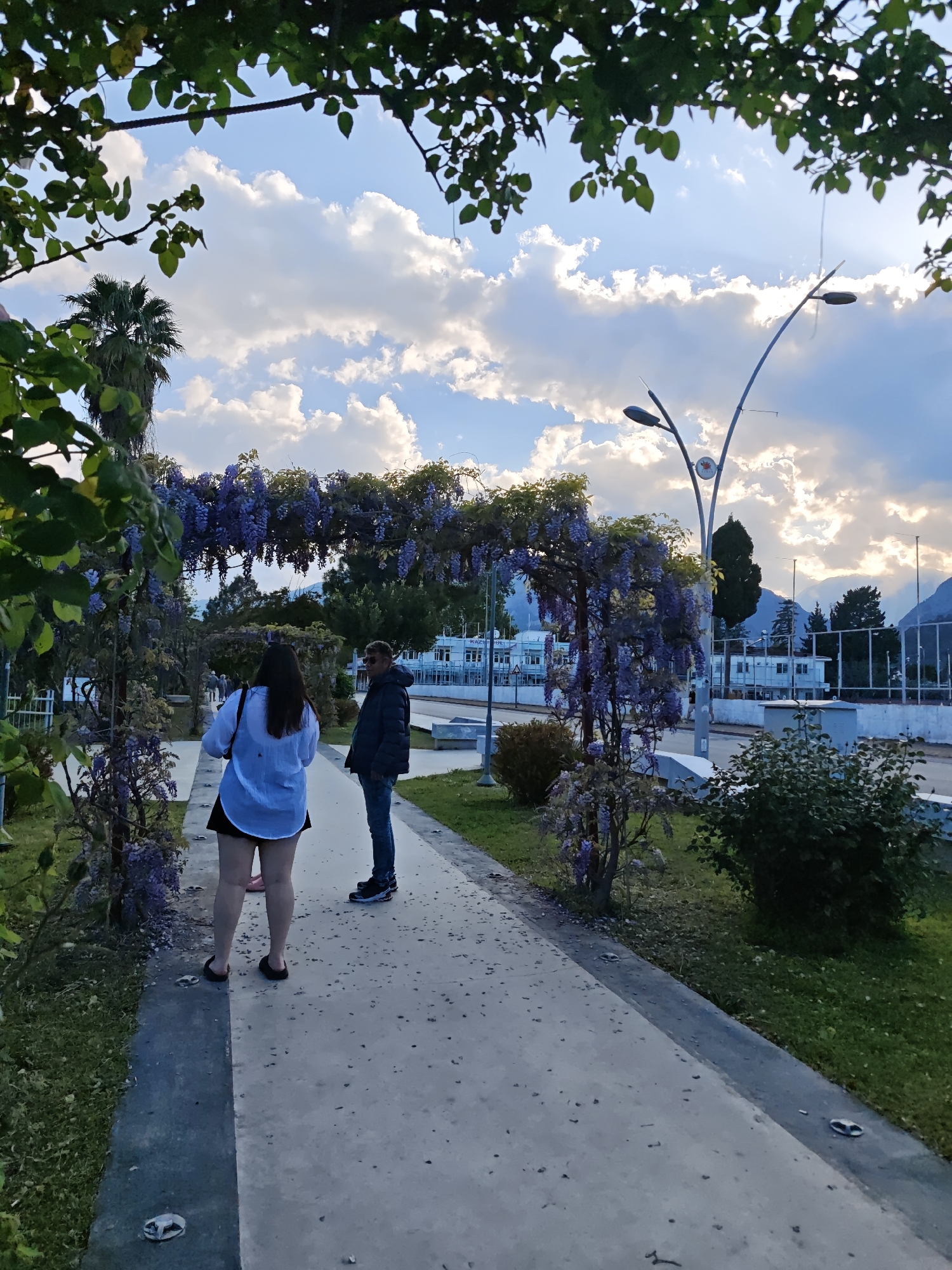 Two people stand on a concrete path under an arbor covered in purple flowers in a park in Kemer, Turkey. A woman with long dark hair wears a light blue shirt and black shorts. A man, wearing a dark jacket and jeans, stands beside her, looking towards the right. Behind them, a white building is visible, along with mountains in the distance. The sky is filled with clouds and the sun peeks through. A lamppost and various plants and trees line the path. Litter is visible along the side of the path.