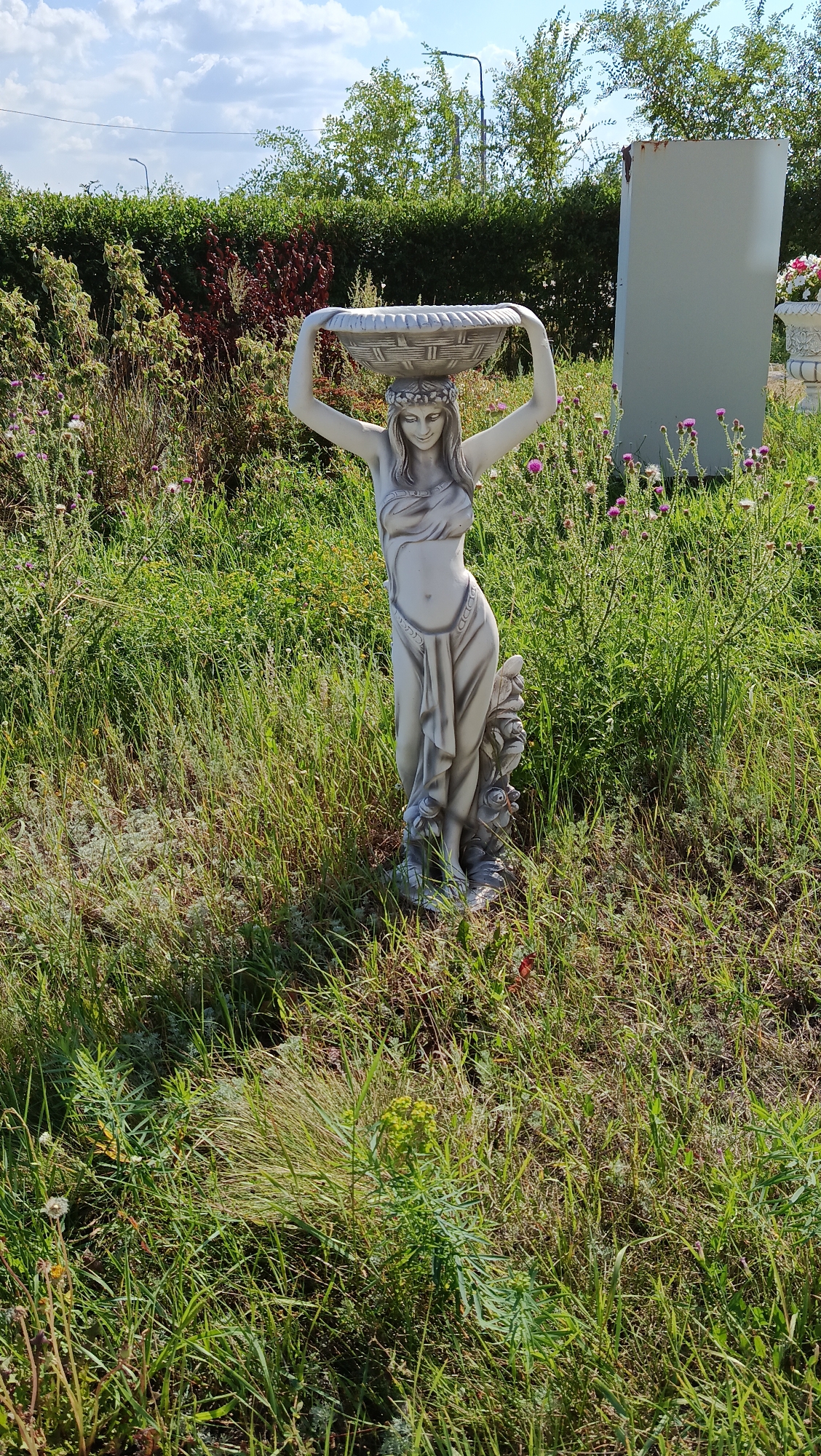 An outdoor, eye-level, full shot shows a concrete statue of a woman with her arms raised above her head, supporting a basket. The statue is light grey and depicts a female figure with long, flowing hair adorned with a flower crown. She is wearing a draped garment that exposes her midriff. Her body is slender and posed in a classical style. The statue stands in a grassy, unkempt area with tall weeds and small purple flowers. In the background, a dense green hedge runs horizontally across the frame. To the right of the statue, a tall, rectangular, light grey structure is visible, possibly a memorial or marker, with a large planter containing white and pink flowers to its right. The sky above is blue with scattered white clouds. The overall impression is of a decorative statue placed in a naturalistic, overgrown garden setting.