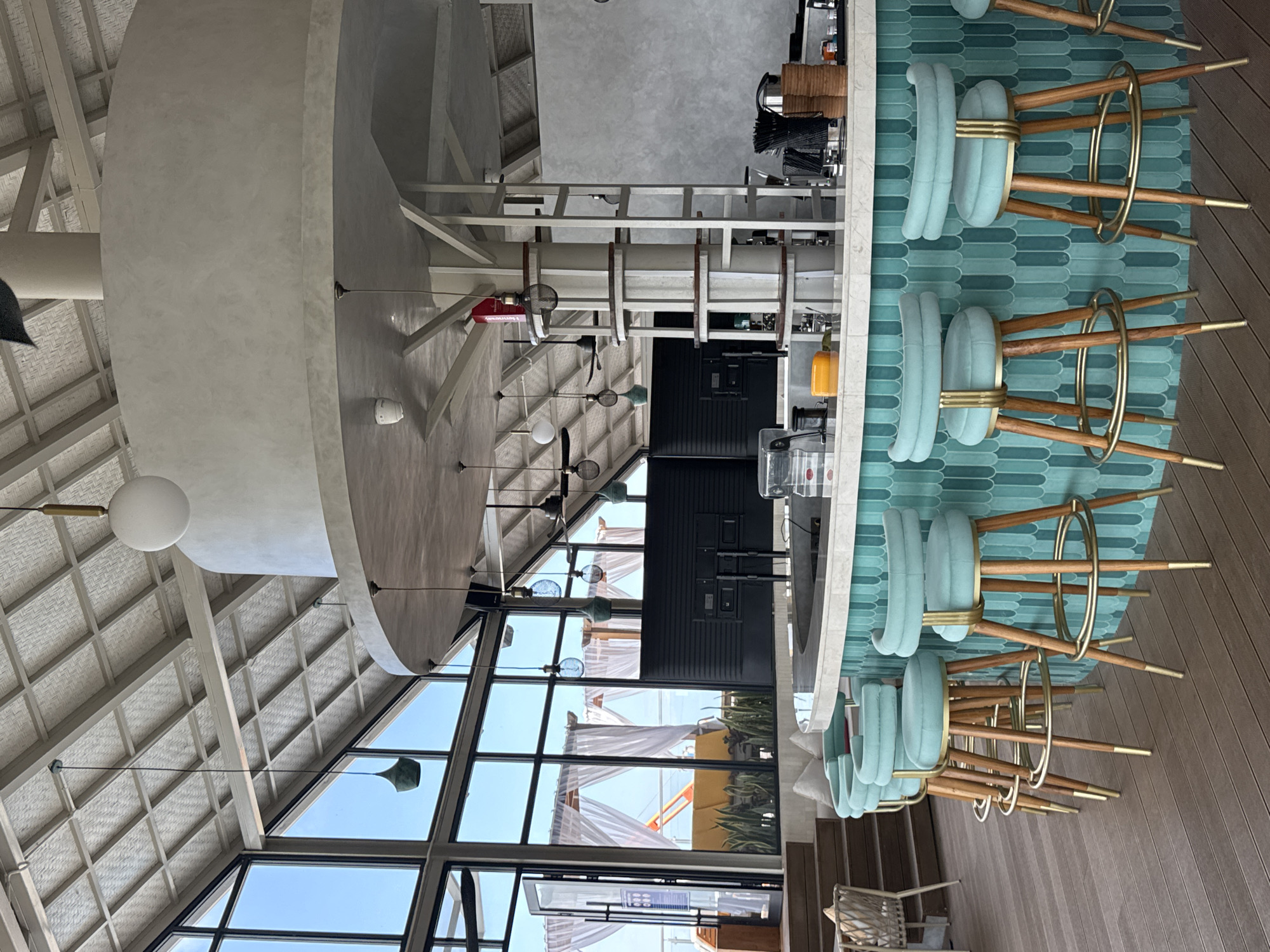 The image depicts a bar area in North Kuta, Indonesia. In the foreground, several light blue bar stools with wooden legs and gold accents are positioned around a curved bar. The bar itself has a white countertop and a front panel decorated with teal, elongated oval tiles. Behind the bar, there are two large black screens, bar stools, and various bar equipment. Large windows with a view of outdoor seating areas, white curtains, and the sky are visible in the background. The ceiling is a pitched structure with exposed beams and a textured material. The overall aesthetic suggests a modern, open-air establishment.