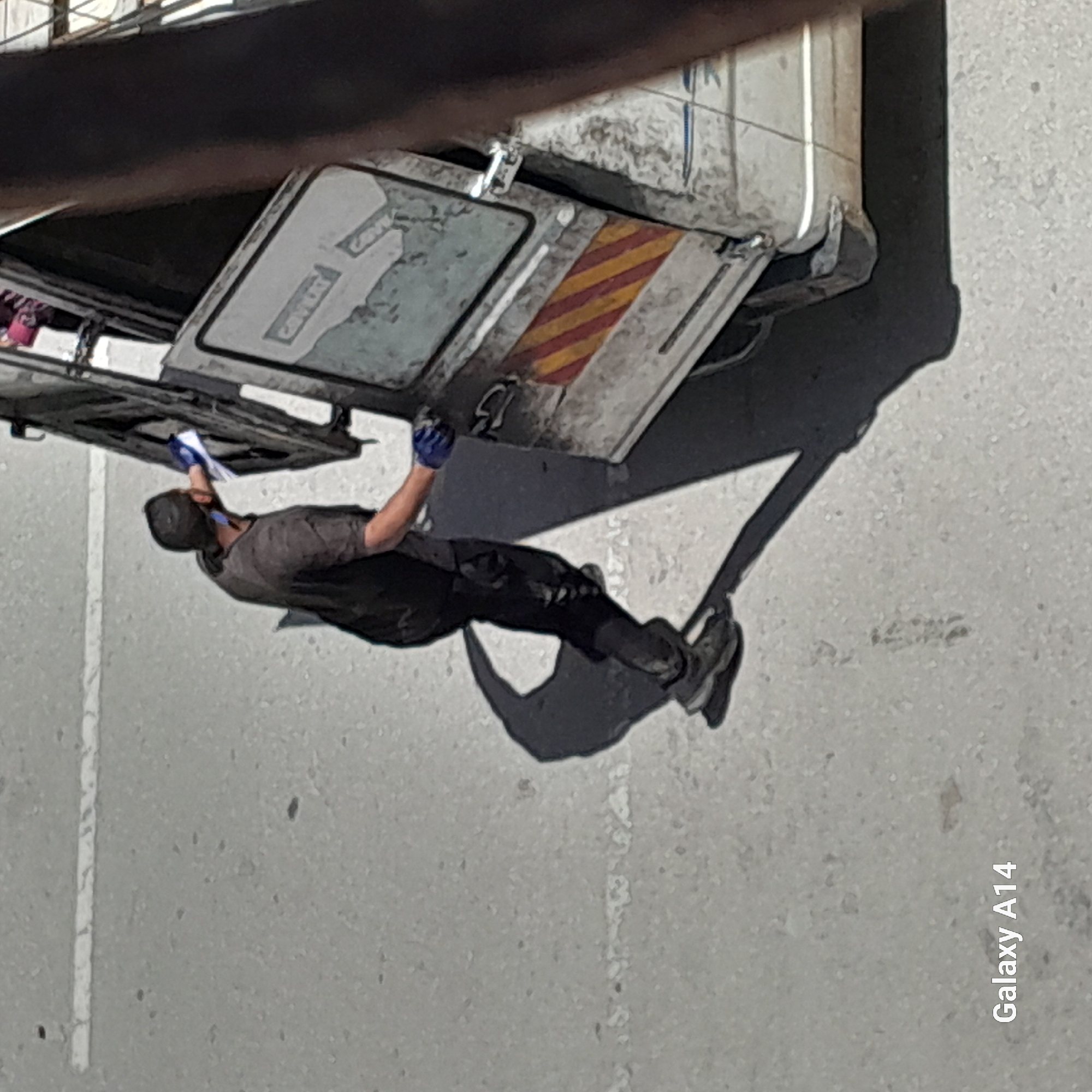 A person is working on the underside of a commercial vehicle. The person is wearing gloves, dark clothing and shoes. The commercial vehicle is light colored with warning striping on the open compartment door. The scene appears to be a city street or parking lot in Unknown, Unknown. A painted white line can be seen on the pavement.