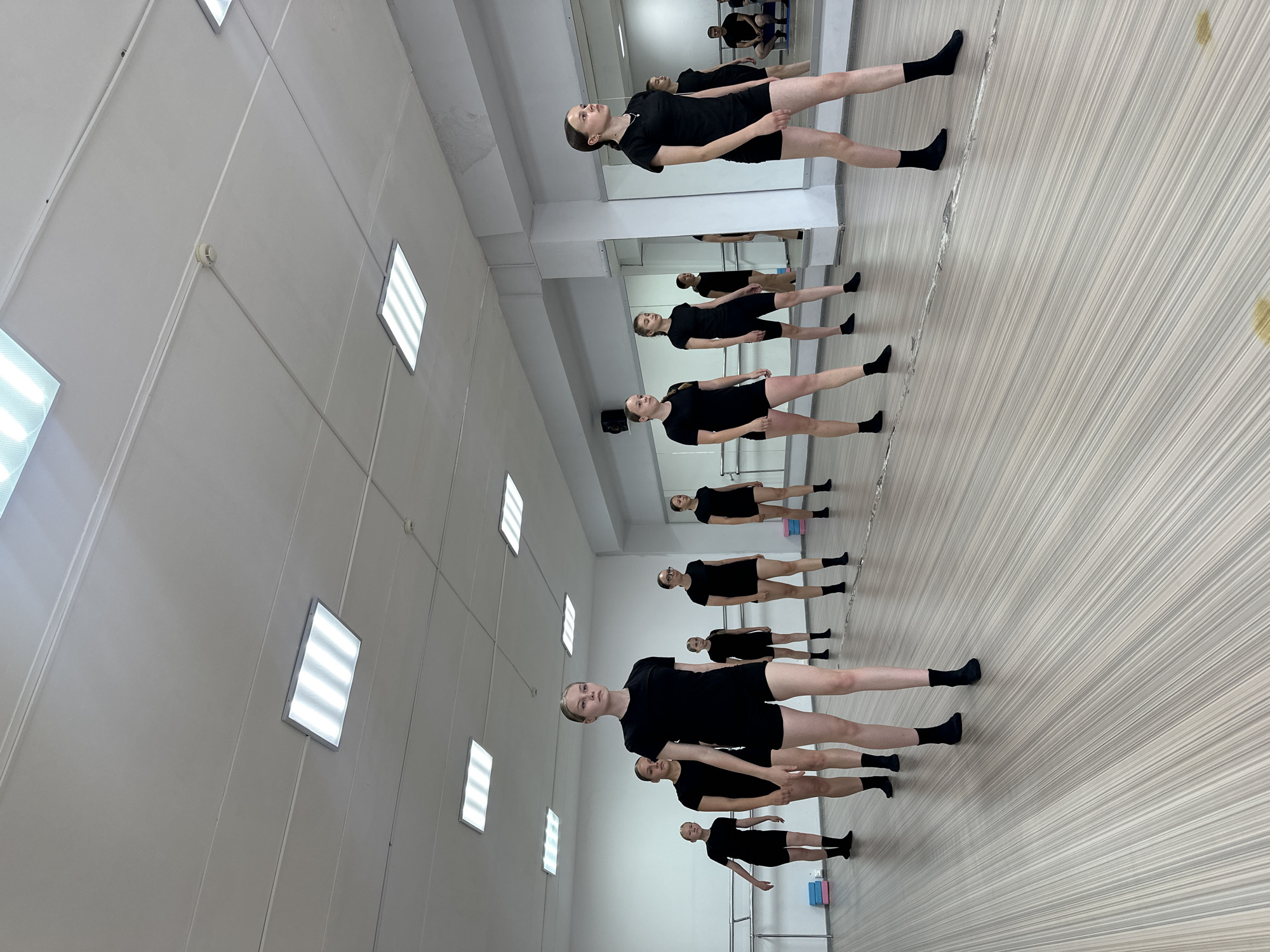 The image shows a group of nine young women in a dance studio. They are all dressed in black leotards and shorts, and black socks. They are arranged in two rows, facing forward, in what appears to be a dance class. The studio has a wooden floor, a mirrored wall, and a ballet barre along one wall. There are also fluorescent lights on the ceiling. In the background, there are some small, colorful blocks on the floor, and in a mirror reflection on the right side of the image, a person is sitting on a bench. The studio is clean and well-lit. It is not possible to determine the specific location in Votkinsk, Russia from the image alone, but it is likely a dance school or studio.