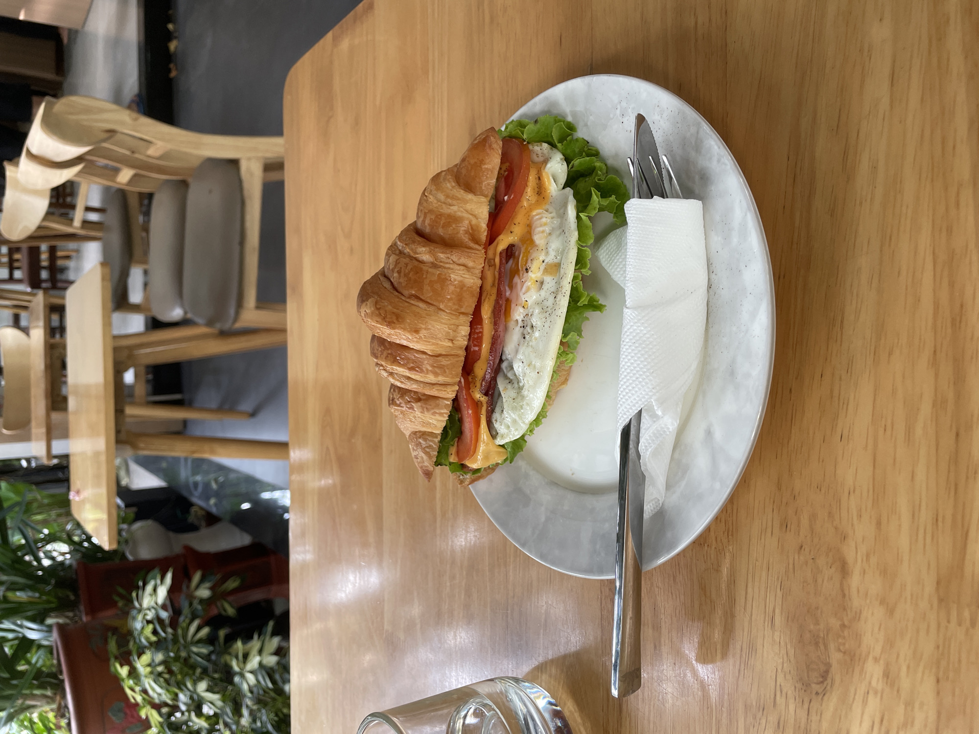 A croissant sandwich rests on a white plate, which is placed on a wooden table. The sandwich is filled with lettuce, tomato, cheese, bacon, and a fried egg. A fork and knife are placed on the plate, alongside a white napkin. A glass of water is to the left of the plate. In the background, there are chairs and tables, suggesting the location is a cafe or restaurant. The plants in the background indicate it's an indoor setting with some natural elements. The overall scene captures a meal in a cafe in Da Nang, Vietnam, focusing on the food and the dining environment.