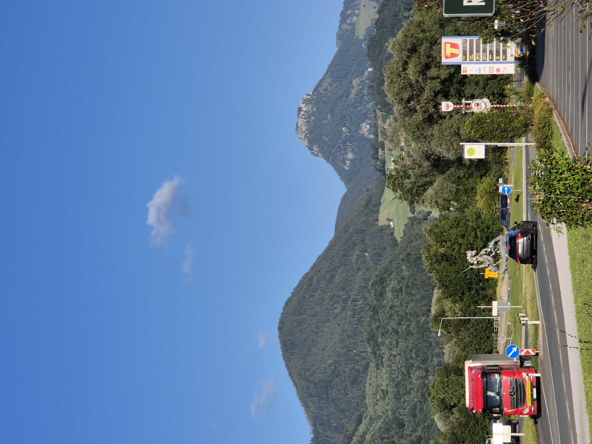 A bright, clear blue sky is visible above a mountainous landscape. The foreground features a road with several vehicles. A large red truck with a white trailer is on the left side of the road, turning. A dark SUV is in the right lane, moving away from the camera. There are traffic signs and a sculpture of a knight on a horse fighting a dragon in the background, situated on a roundabout. A gas station with price displays and signage for "T" fuel is also visible. Trees and green hills form the midground, leading to the imposing, tree-covered mountains in the distance. The overall scene depicts a roadside environment in a mountainous region, under a sunny sky.