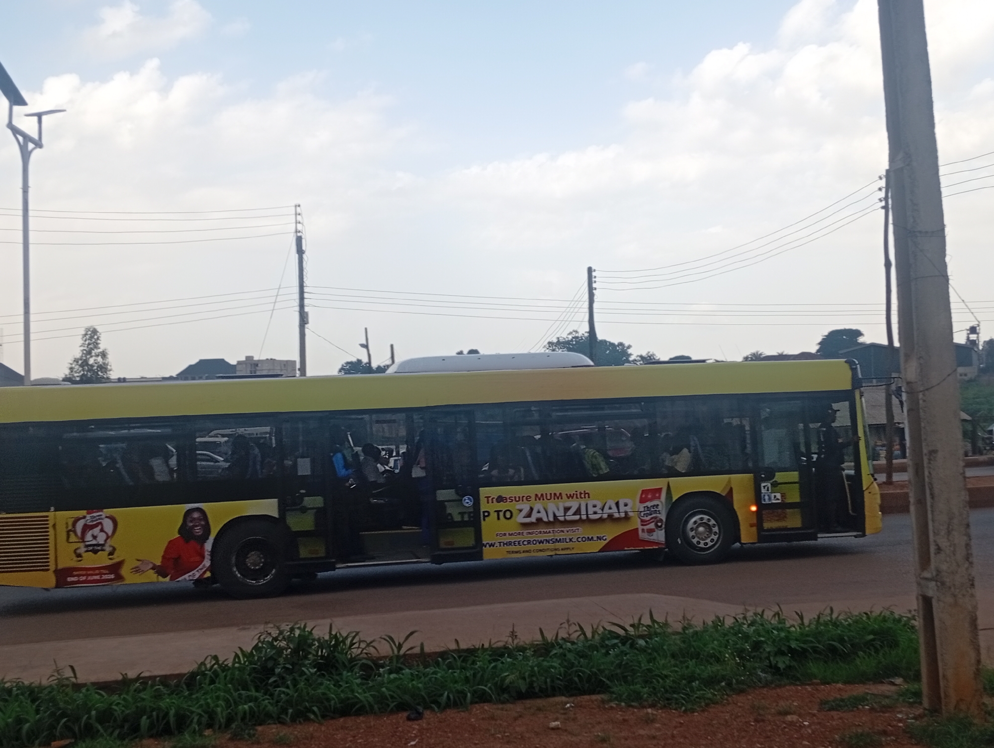 The image shows a side view of a yellow public transport bus driving on a paved road in the city of Bukuru, Nigeria. The bus has tinted windows, and several passengers are visible inside. Advertisements are displayed on the side of the bus. One advertisement features a woman wearing a sash and the text "QUEEN OF THE." Another advertisement promotes a trip to Zanzibar, with the text "Treasure MUM with P TO ZANZIBAR" and the website "WWW.THREECROWNSMILK.COM.NG". Power lines and utility poles are visible in the background, along with some buildings and trees. The ground in the foreground is covered with red dirt and patches of green grass. The sky is partly cloudy.