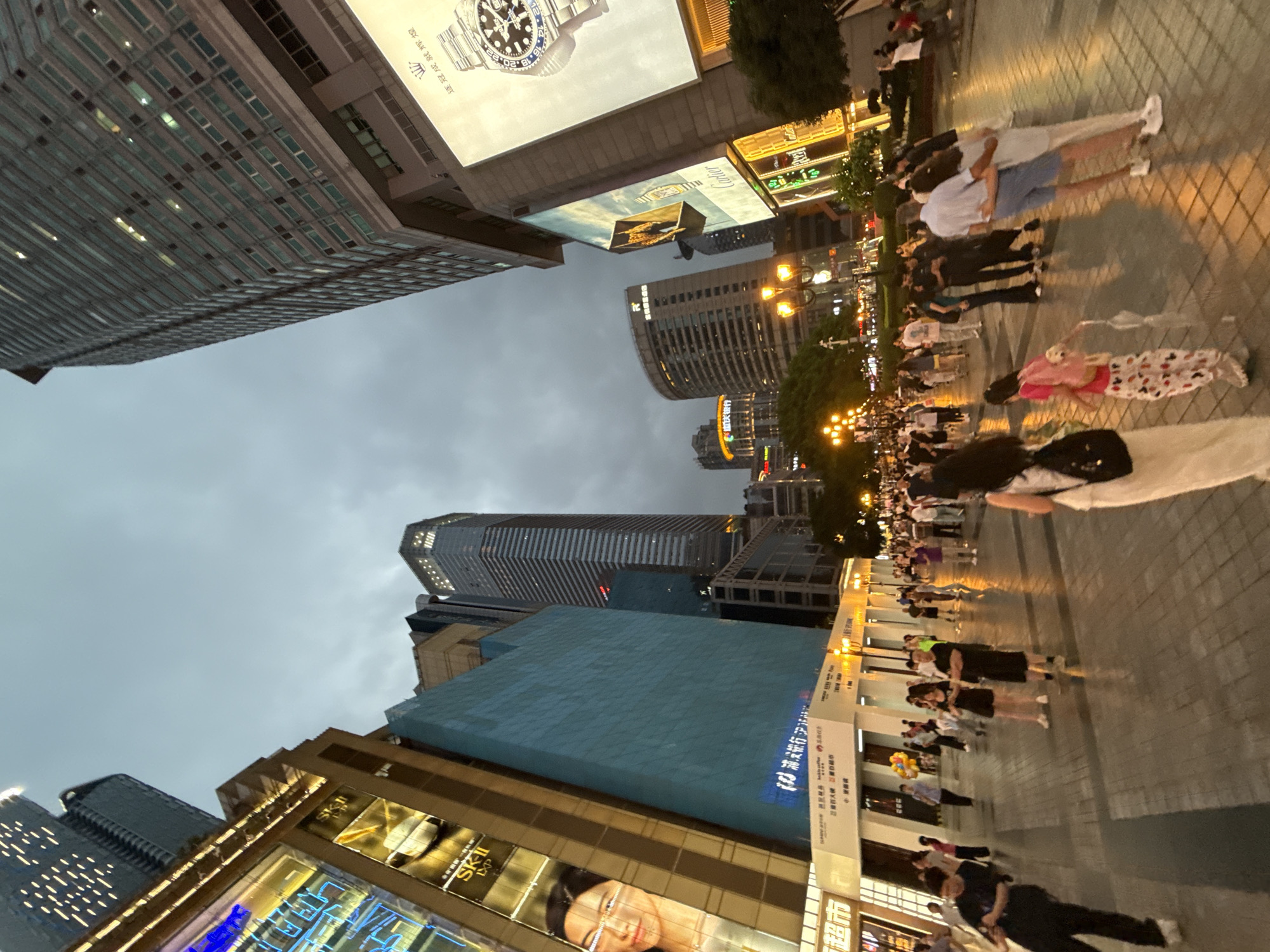 This is a low-angle, full-shot, eye-level photograph of a bustling pedestrian street at dusk in Yuzhong District, China. The scene is dominated by towering, modern skyscrapers with illuminated signage and windows, characteristic of an urban commercial area. In the foreground, a paved pedestrian walkway is filled with a diverse crowd of people walking, standing, and interacting. The lighting suggests it is evening, with artificial lights from buildings and street lamps casting a warm glow on the scene, creating reflections on the wet pavement.
Several large buildings are visible, including a skyscraper with many windows on the right, and a distinct building with a curved facade on the left, displaying large advertisements. One prominent advertisement features a close-up of a woman's face and the text "SK-II". Another large advertisement on the right showcases a Rolex watch. Smaller shops and businesses are visible at street level, with illuminated signs.
The people in the image are dressed in casual summer attire. They are engaged in various activities: some are walking in groups, others are standing and talking, and a few are holding hands, suggesting social gatherings or leisure time. A young girl in a pink backpack is walking with an umbrella. The overall atmosphere appears lively and active, typical of a popular urban destination in the evening. The sky is overcast, contributing to the dusk lighting conditions.