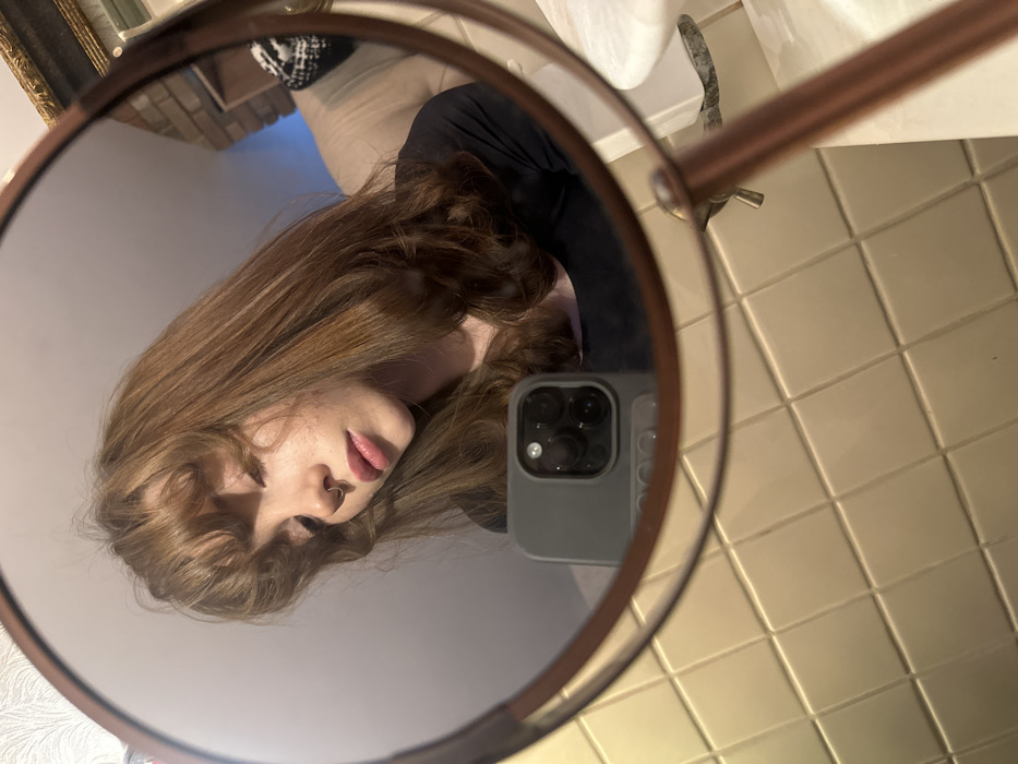 A mirror selfie is captured, showing the reflection of a female individual and a bathroom interior. The primary subject, a female with shoulder-length reddish-brown hair and bangs, a septum piercing, and wearing a black scoop-neck top, is reflected with her head tilted down and to the left, eyes downcast, and lips slightly parted. She holds a grey-cased smartphone with a multi-lens camera visible in the lower center of the reflection. Behind her, a partial reflection of a second individual wearing a light-colored top and a black-and-white patterned head covering is visible to the upper right. The background of the reflection includes a tiled wall with light-colored square tiles, a white sink basin with a marbled countertop, and a chrome faucet. Exposed brickwork and a partially visible gold-framed object are also present in the upper right background. The mirror itself has a copper-toned frame and a visible stand. The location is an indoor bathroom in the city of Unknown, Unknown.