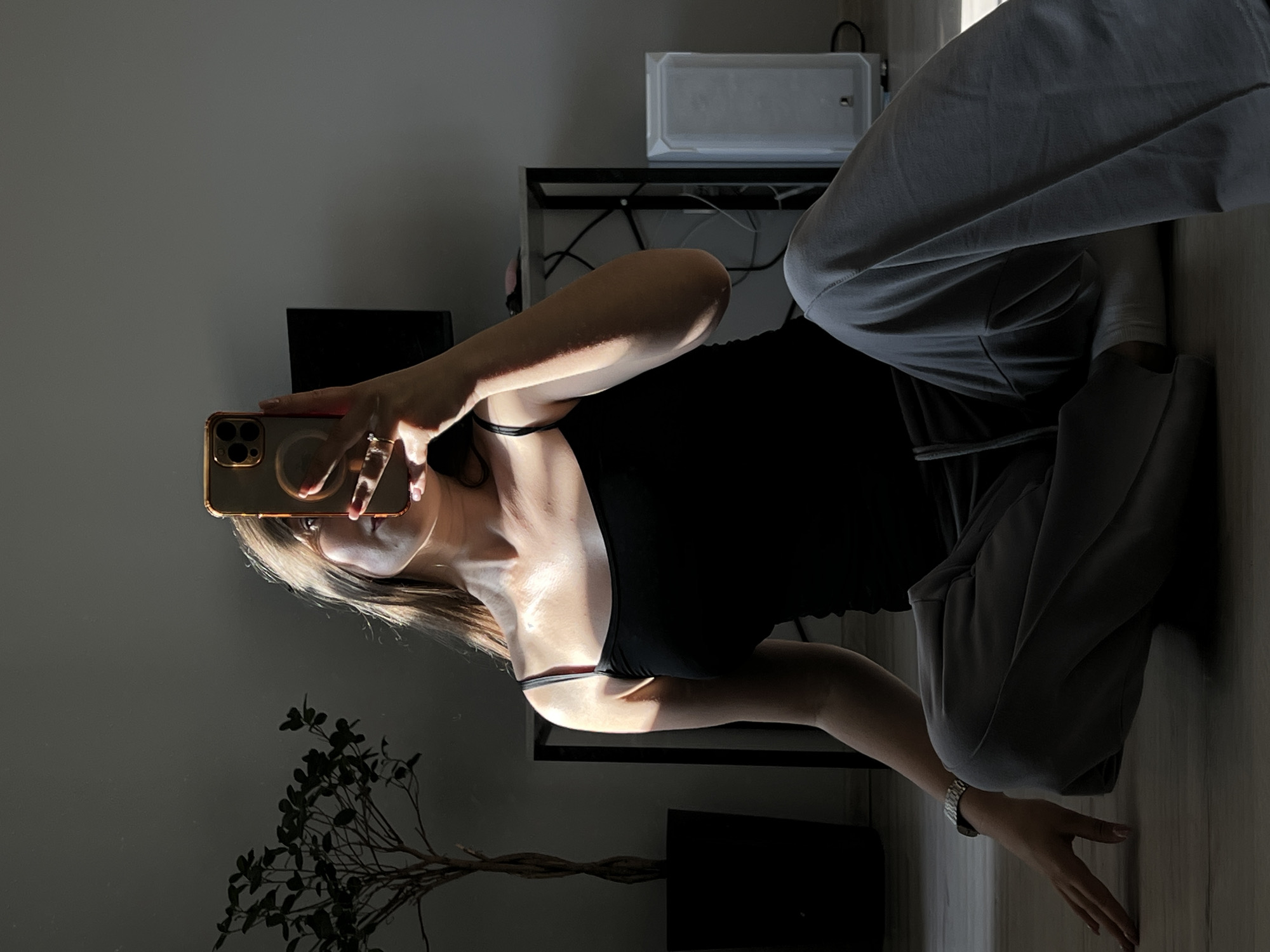A woman is sitting on the floor in a dimly lit room, taking a selfie with her phone. She is wearing a black tank top and gray sweatpants. A plant in a black pot is visible to her left, and a white air purifier and a desk with a television are in the background. The room appears to be a residential interior in Ternopil, Ukraine, as suggested by the mention of the city. The lighting suggests it is daytime, with sunlight streaming in from a window.