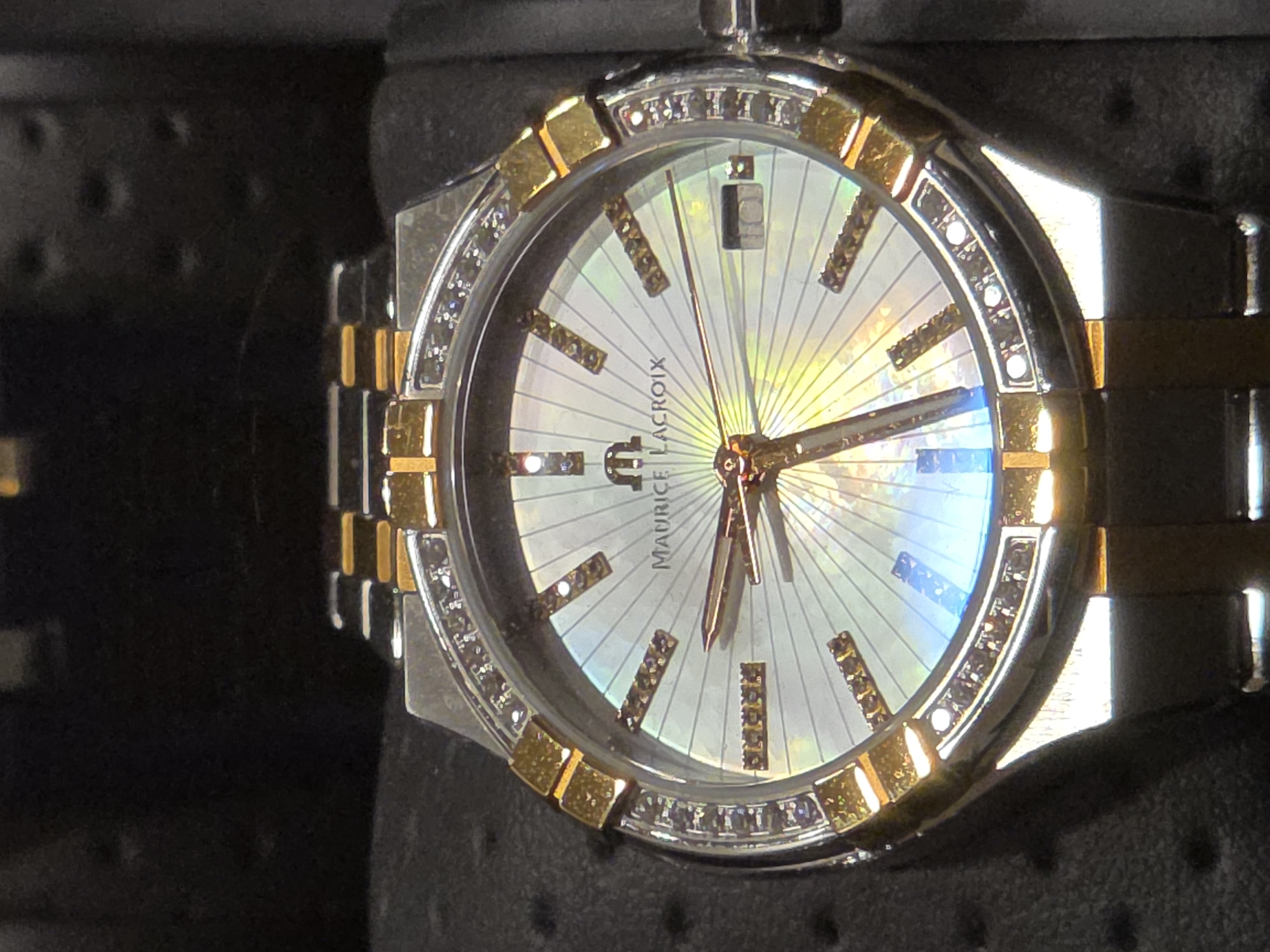 A close-up shot shows a Maurice Lacroix watch with a mother-of-pearl dial. The watch has a two-tone band with silver and gold links and a bezel decorated with diamonds. The watch face features diamond hour markers, silver hands, and a date display at the 12 o'clock position. The watch is positioned against a dark, textured background.
The location is not discernible from the image and there is no information about it being in Warsaw, Poland.
