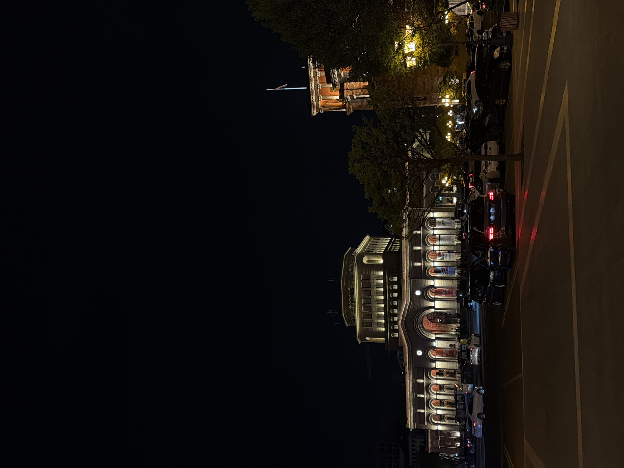 The image captures a vibrant night scene in an urban setting, likely Yerevan, Armenia, as indicated by the context. The photograph is taken from a slightly elevated perspective, looking across a street or square.
The most prominent subjects are two large, ornate buildings illuminated by warm artificial lights. In the foreground, a grand, multi-story building, possibly in a classical or historic architectural style, dominates the right side of the frame. Its facade features several large arched entrances or windows at street level, and rows of smaller, illuminated windows on upper floors. The building's detailed architecture, with columns and decorative elements, is highlighted by the strategic lighting.
Further back and to the left, another substantial building with a distinctive tower or spire is visible, also illuminated against the dark sky. A flag, appearing to be the Armenian flag (red, blue, orange), is visible atop this structure, providing a key geographical cue.
Below the buildings, a street or parking area is visible, with several cars parked along the curb. Some vehicles display glowing red tail lights. Dark silhouettes of trees are interspersed between the buildings and along the street, their foliage subtly illuminated by the ambient lights. The paved road surface features clear yellow markings, including linear stripes and a large chevron or arrow pattern.
The time of day is definitively night, with a completely dark sky creating a strong contrast with the brightly lit architectural elements. The overall atmosphere is calm and urban, showcasing the city's illuminated nighttime appeal. No discernible human figures are clearly visible, and there is no legible text or signage.