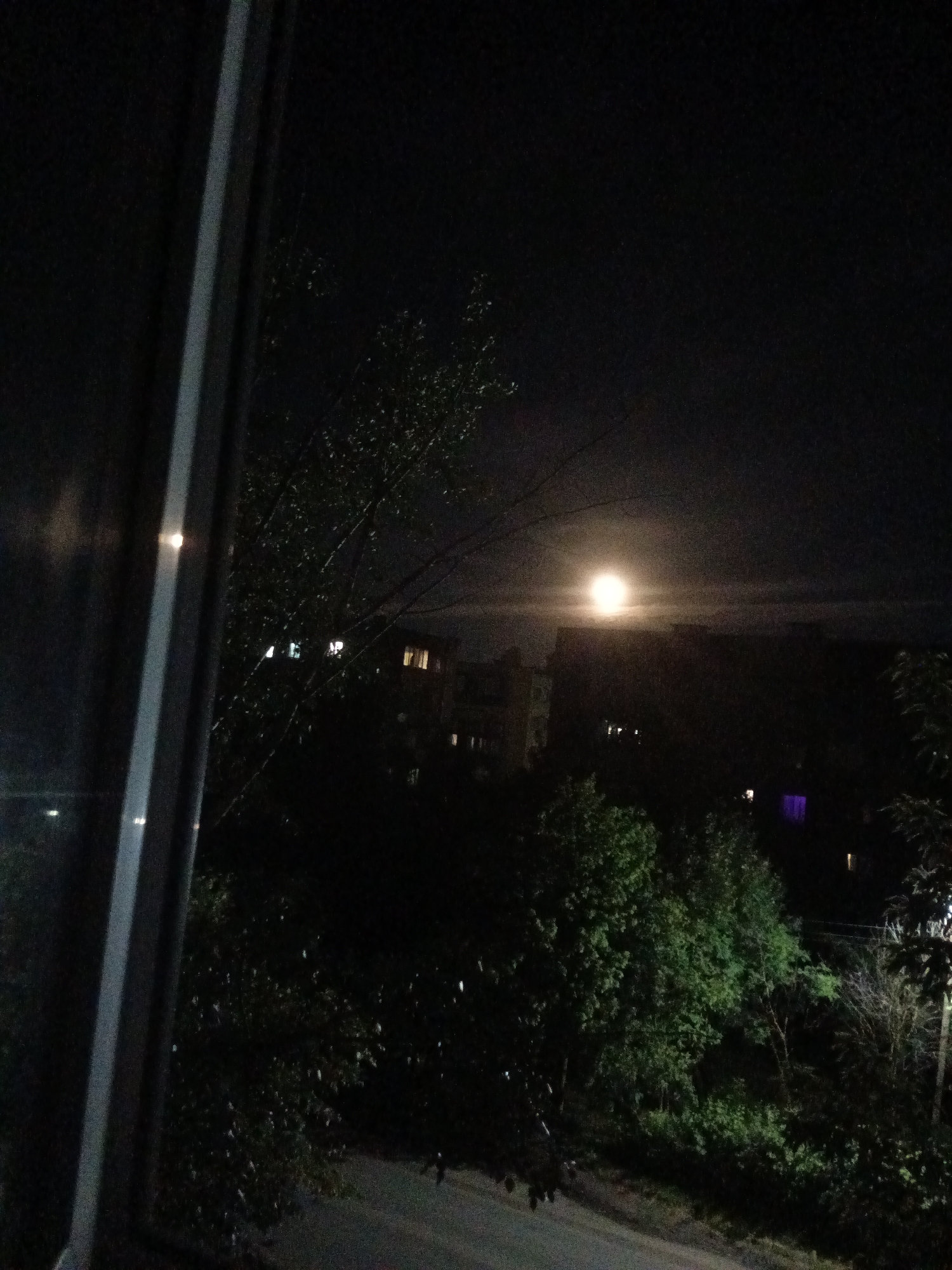 An outdoor night scene in Kuznetsk, Russia, is captured through a window frame, which partially obscures the view on the left. A bright, full moon is visible in the upper central part of the sky, casting a soft glow. Silhouetted trees with visible branches and foliage dominate the foreground and middle ground. In the background, several multi-story buildings with illuminated windows are discernible. The overall atmosphere is dark and nocturnal, with the moon and building lights providing the primary illumination. A road is visible in the lower part of the image, appearing wet and reflecting some light.