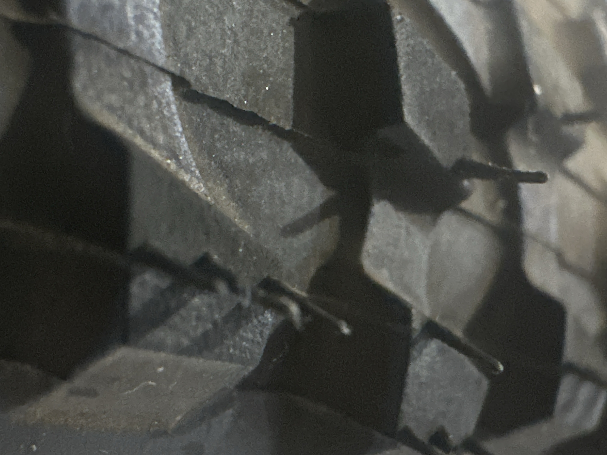 The image shows a close-up, low-angle view of the tread pattern on a tire. The tire appears to be dark in color, likely black rubber, and the tread blocks are deeply grooved and angular, suggesting it is a heavy-duty or off-road tire. Some small, light-colored particles are scattered on the surface of the tire. The lighting is dim, casting shadows that highlight the texture and depth of the tire's design. There are no people or discernible environmental cues to indicate the time of day, weather, or a specific location beyond what can be inferred from the object itself. The image focuses solely on the intricate details of the tire tread. No visible text is present.