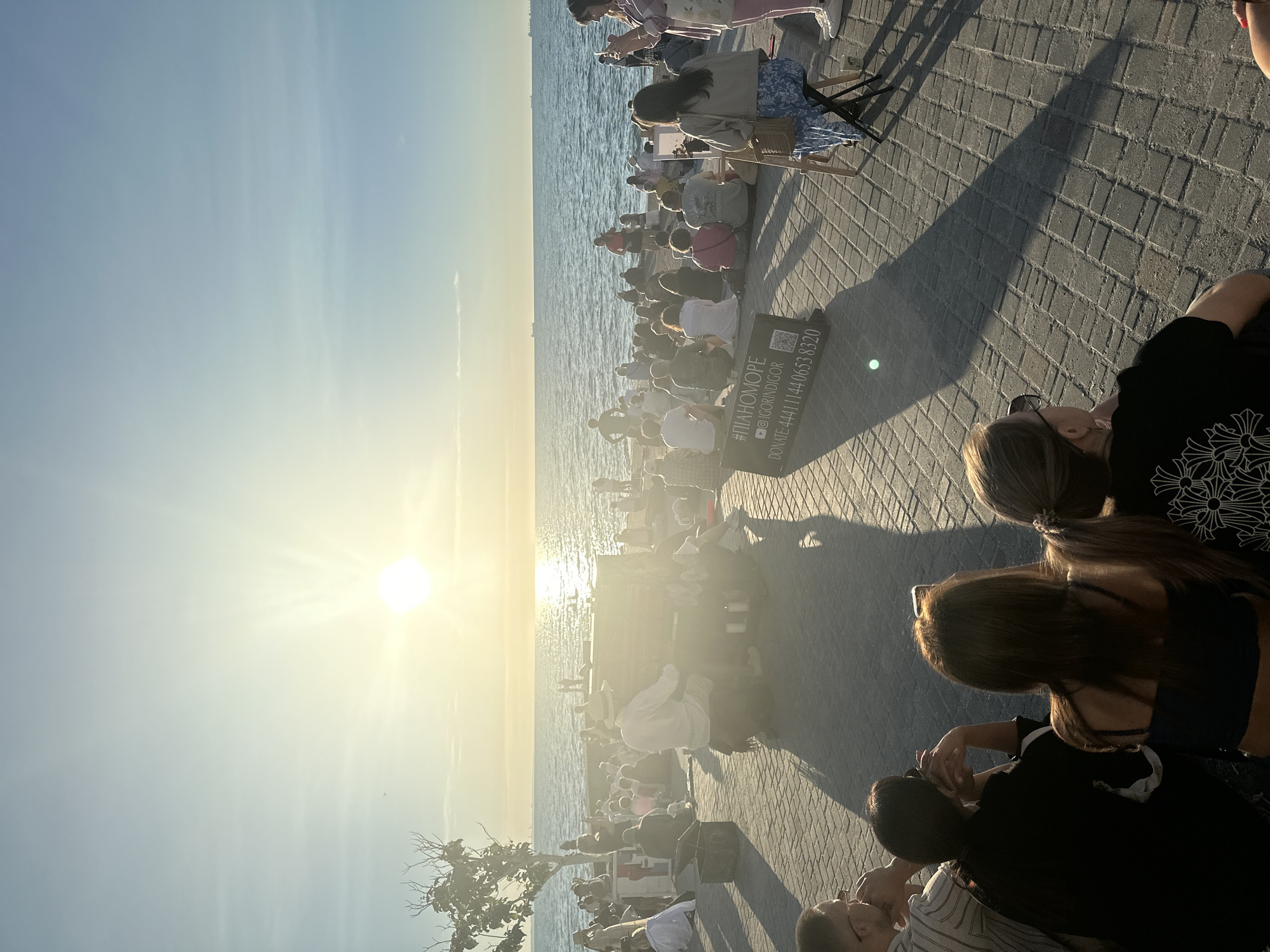 The image depicts a scene on a seaside promenade in Odesa, Ukraine, during what appears to be sunset. The sun is low in the sky, casting a warm glow and creating long shadows on the paved walkway. A crowd of people are gathered, many sitting on the ground or on low walls, facing the water.
In the foreground, the backs of several individuals are visible, suggesting they are part of the audience. Further back, a man wearing a hat is seated at a piano, playing for the assembled crowd. A woman is seated to the right of the piano, appearing to sketch or paint on an easel. A sign with Russian text, including a hashtag and a phone number for donations, is placed on the ground near the crowd.
The sea stretches out to the horizon, with a few ships visible in the distance. The sky is clear with a gradient of blue and yellow hues from the setting sun. The promenade is paved with brick-like stones, and a few sparse trees are present along the waterfront. The overall atmosphere suggests a public gathering for an outdoor performance.