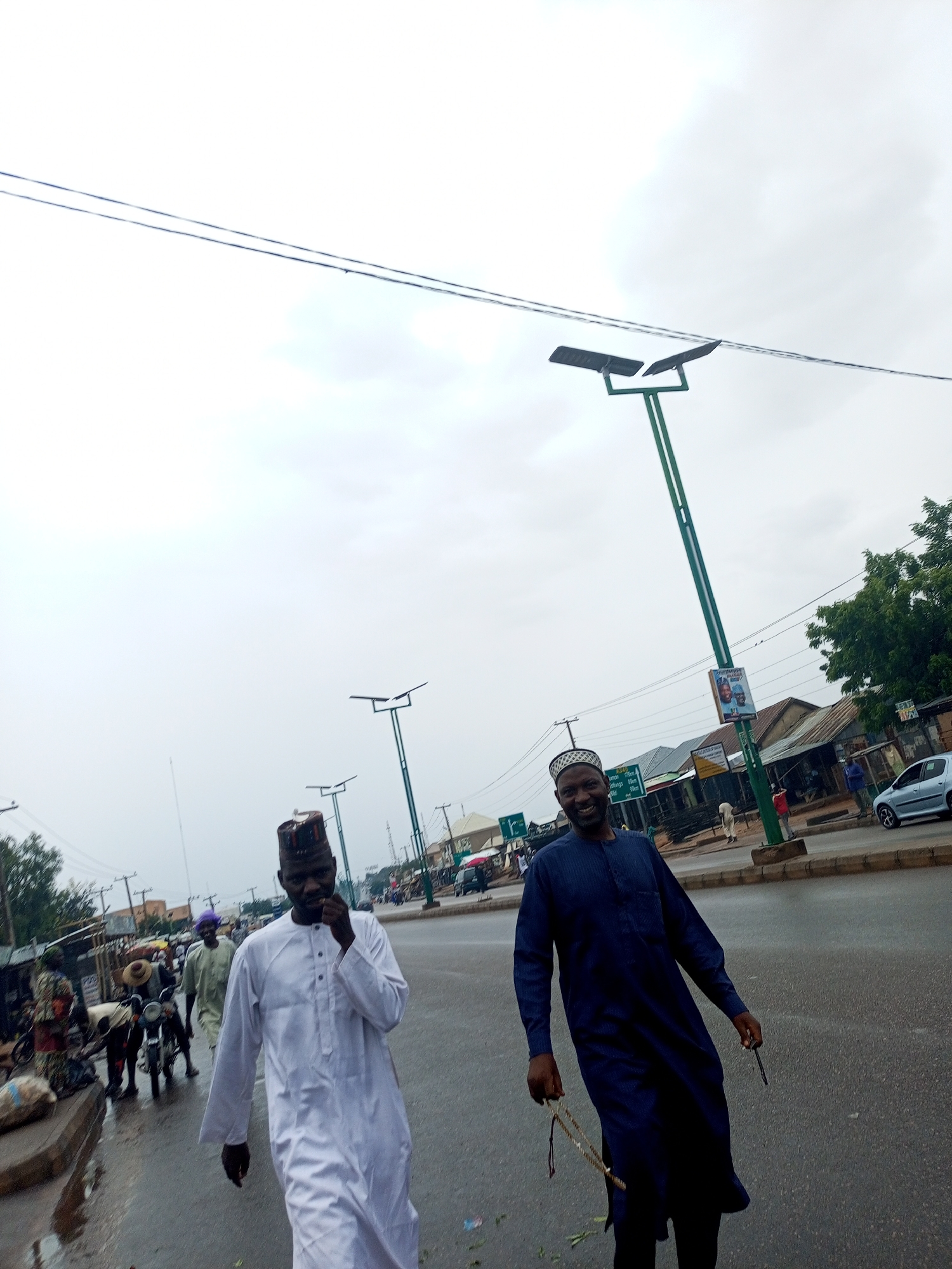 The image captures a vibrant street scene on what appears to be an overcast day, possibly after rain, given the damp road surface. Two men are prominently featured walking along the street. The man in the foreground on the right wears a dark blue traditional kaftan and a white skullcap (kufi), smiling broadly towards the viewer while holding what looks like a string of prayer beads (tasbih) and another small object. The man to his left, slightly behind, is dressed in a white traditional kaftan and a patterned cap, with his right hand near his mouth, his expression less clear.
In the background, the street is busy with other pedestrians and motorcycles. Buildings, likely shops or stalls, line both sides of the road. Several green-poled solar-powered streetlights are visible, extending into the distance. Overhead, utility wires crisscross the frame against the bright, cloudy sky. A silver car is parked on the right side of the road further down.
Notable details include a large green directional road sign on the left, which lists several Nigerian cities and their distances, including "Gombe 2km," "Potiskum 49km," "Bauchi 167km," and "Jalingo 287km," indicating the location is likely in northeastern Nigeria. Another sign, possibly a political campaign poster featuring two faces, is attached to one of the streetlight poles. The overall atmosphere suggests a typical day in a bustling Nigerian town or city.
