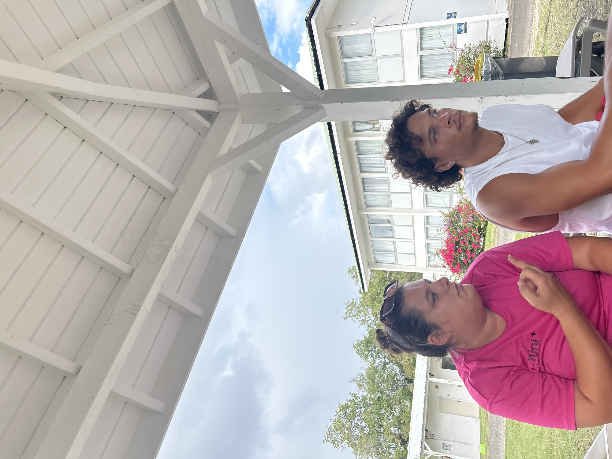 Two individuals, a man and a woman, are seated outdoors under a white wooden structure, likely a gazebo or covered area. The man, with dark curly hair, is wearing a white sleeveless shirt and a silver necklace. He is looking upward, away from the camera. The woman, wearing a pink t-shirt with a logo, has dark hair pulled back and sunglasses perched on her head. She is looking upwards and pointing with her right hand. The background includes white buildings with windows, green trees, flowering plants, and a view of the sky with clouds. The location may be in Sainte-Anne, France, as specified in the instructions.