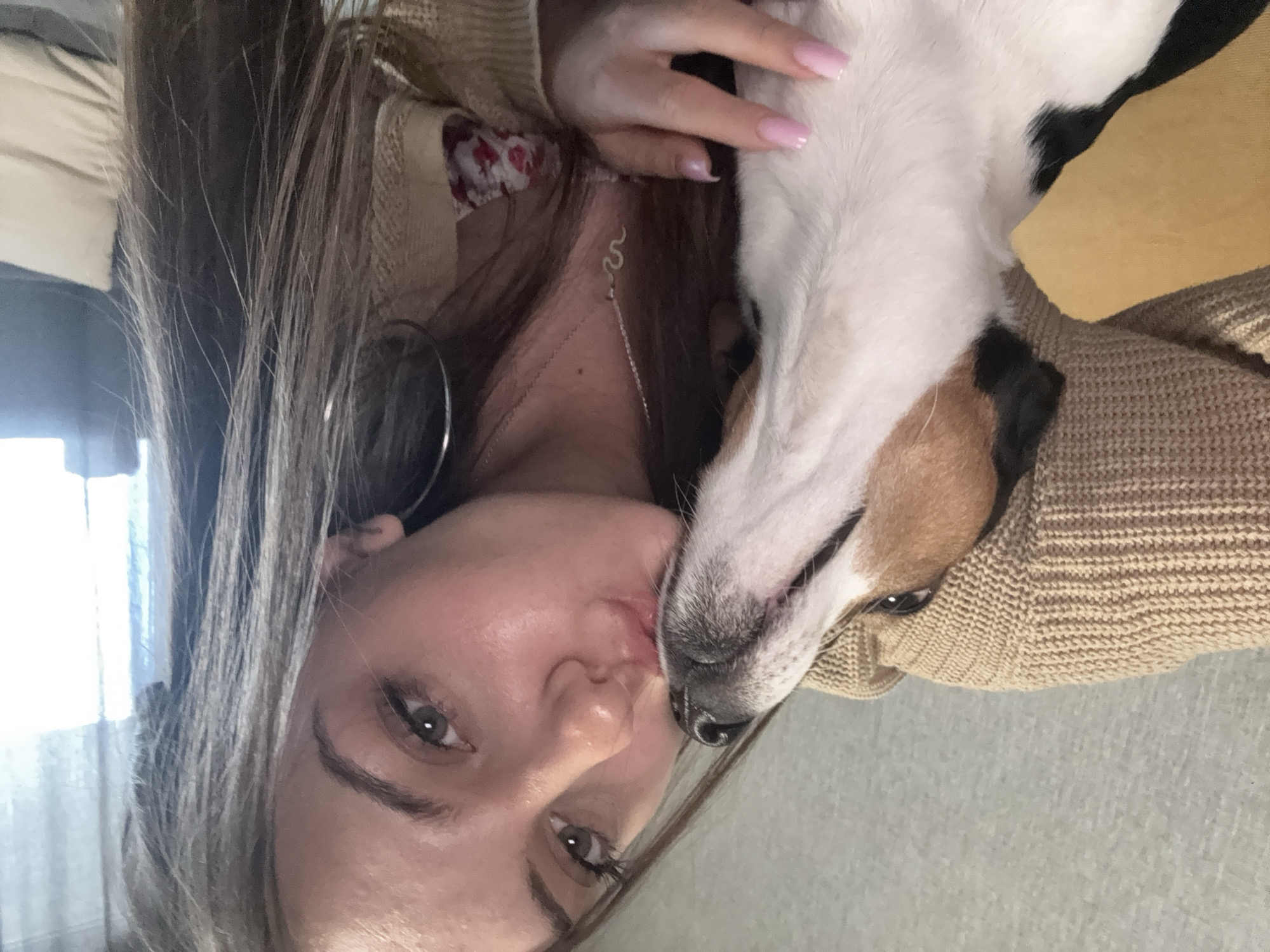 A woman is lying down and facing the camera. She is wearing a tan sweater, a white shirt with a red floral print, and a silver snake necklace. She has long brown hair, silver hoop earrings, and pink manicured nails. Her face is close to the camera. A dog with white, tan, and black fur is lying on top of her and partially covering her face. The dog is looking towards the camera. The background is blurry and gray, suggesting the scene may be indoors on a couch. There is no information in the image to indicate that this was taken in Sofia, Bulgaria.