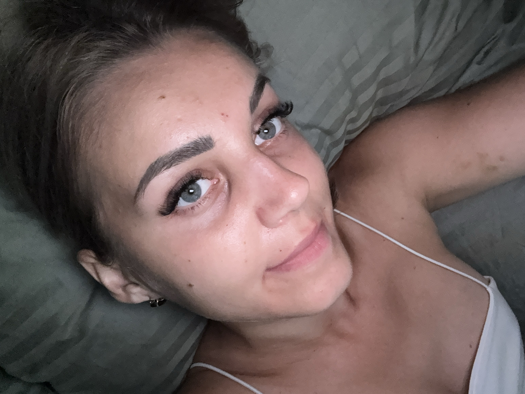 A close-up, high-angle selfie shows a young woman lying on a striped green blanket. She has light skin, grey eyes, dark eyebrows, and false eyelashes. Her lips are slightly parted, revealing a hint of pink lipstick. A thin white strap of a camisole is visible on her left shoulder. A small dark earring adorns her left ear. The lighting is dim, creating shadows on her face. The image is taken indoors, likely in a bedroom, in Yuzhno-Sakhalinsk, Russia.