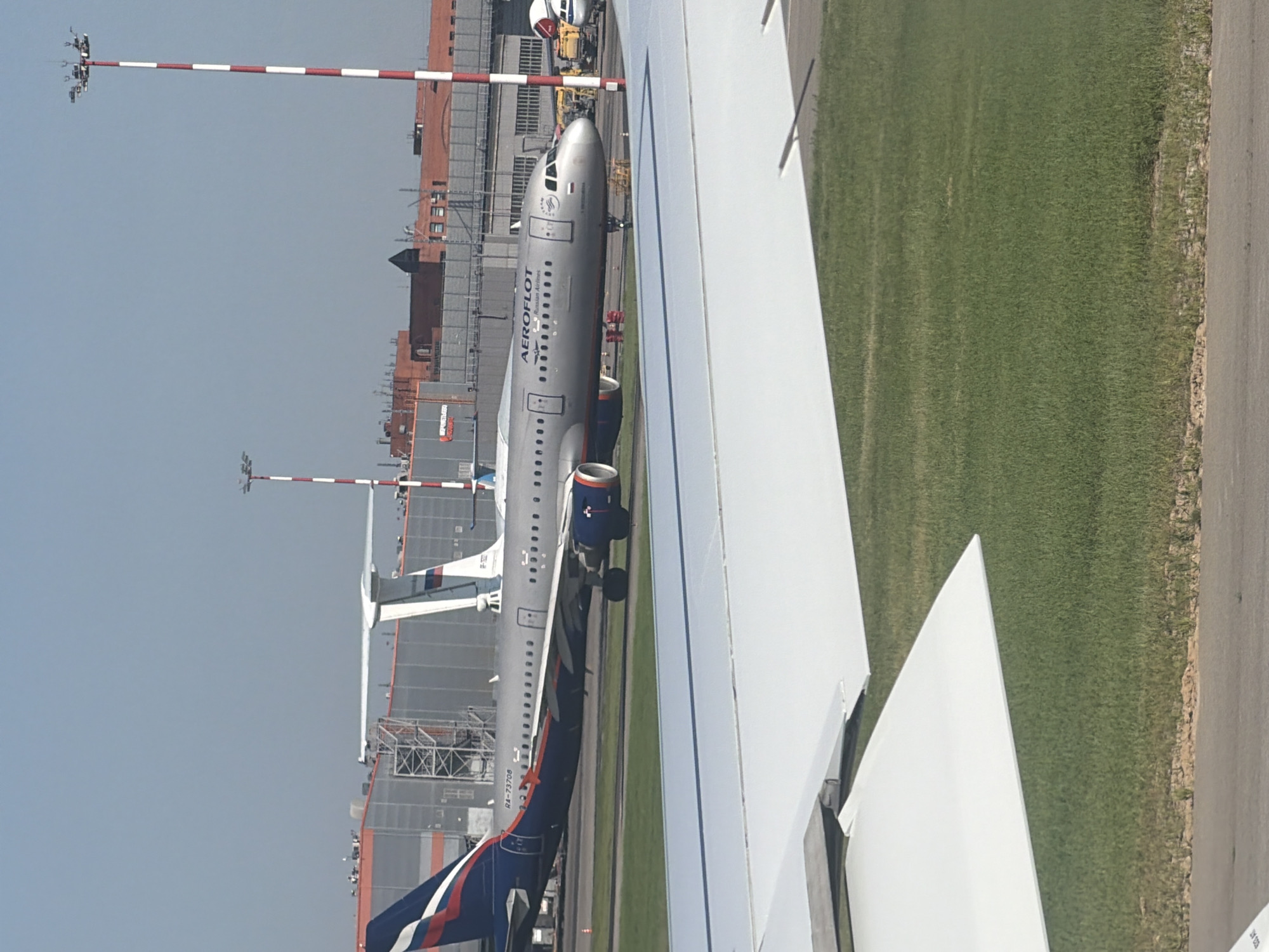 An Aeroflot airplane, with registration number RA-73708, is parked on the tarmac at Sheremetyevo International Airport in Khimki, Russia. The aircraft is a silver Airbus A320. The plane's tail displays the Aeroflot logo, a red, white, and blue design. The airplane is facing right, with its nose slightly angled towards the viewer. In the background, large industrial buildings and hangars are visible, along with red and white striped light poles. The ground is covered with green grass and asphalt. A portion of a white airplane wing is visible in the foreground, obscuring the lower part of the image. The sky is clear and blue. There are no people visible in the scene.