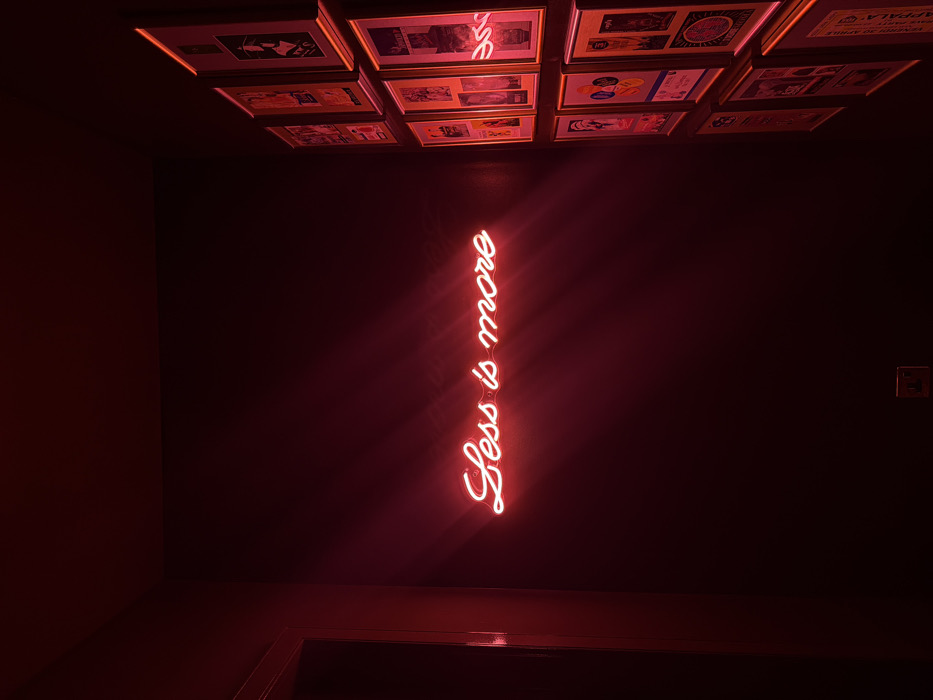 An interior scene in Dubai, United Arab Emirates, depicts a dark room illuminated primarily by a prominent red neon sign. The sign, rendered in a bright cursive script, displays the phrase "Less is more" centered on a dark, unadorned wall. Rays of intense red light emanate from the neon tubing, casting a crimson glow across the immediate surroundings.
To the right of the neon sign, a wall is covered with a grid of approximately eighteen framed artworks or posters, arranged in three vertical columns. These frames, varying slightly in size and orientation, are individually lit by the ambient red light, which obscures specific details of their content, though varied graphic elements are discernible.
On the far left of the image, a vertical section of a dark doorway or passage is visible, reflecting some of the red light. Near the bottom left, an electrical outlet, dark in color, is affixed to the wall. The overall environment is dark and enclosed, with the intense red illumination creating a distinct atmosphere. No individuals are present. The precise location within Dubai is not visually identifiable.