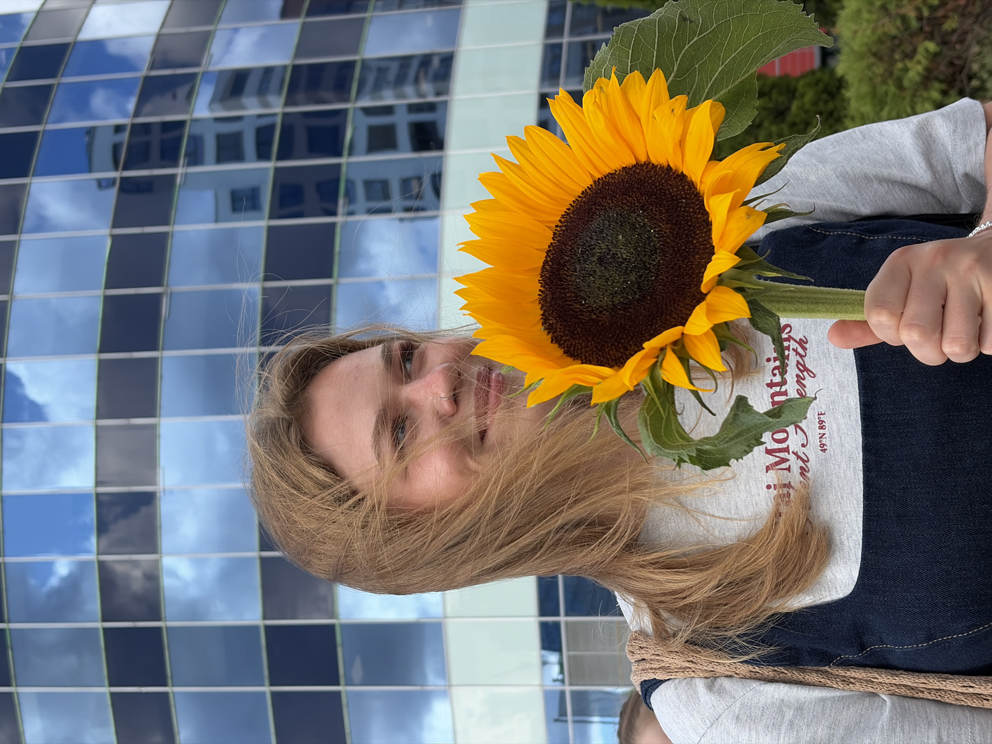 A young woman with blonde hair and a nose ring smiles as she holds a sunflower in front of a modern building with reflective windows. She is wearing a grey t-shirt with text that includes "Mountains" and "49°N 89°E", and a dark denim dress. The building's facade reflects a blue sky with clouds and another building. The scene is outdoors, with greenery visible in the background. The context of Бараўлянскі сельскі Савет, Belarus, is inferred from the image's metadata or associated information, placing the modern architectural structure and surrounding environment within that specific locale.