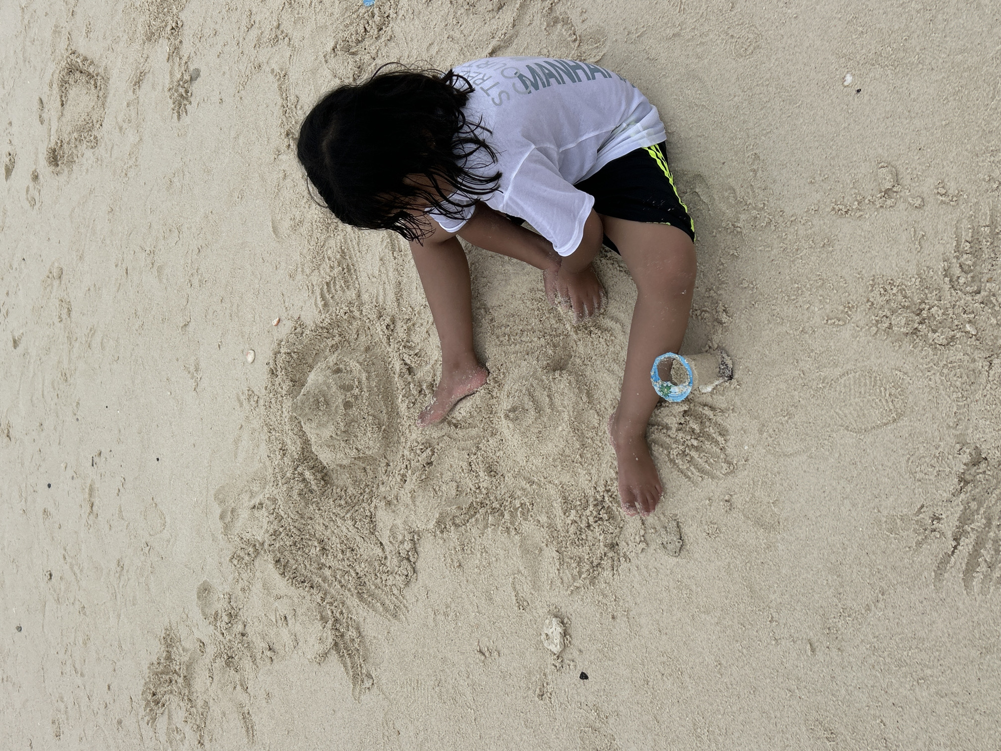 A young child with dark hair is playing in the sand at a beach in Phru Nai, Thailand. The child is wearing a white t-shirt with lettering and black shorts with a neon green stripe. They are sitting on the sand, with their legs extended. A small, partially built sandcastle is visible in front of the child. There is also a blue and white cylindrical object, possibly a bucket or sand toy, near the child's feet. The sand is light in color and appears damp in some areas. The background is entirely composed of sand, with some small shells and debris scattered throughout. The lighting suggests it is daytime, possibly overcast.