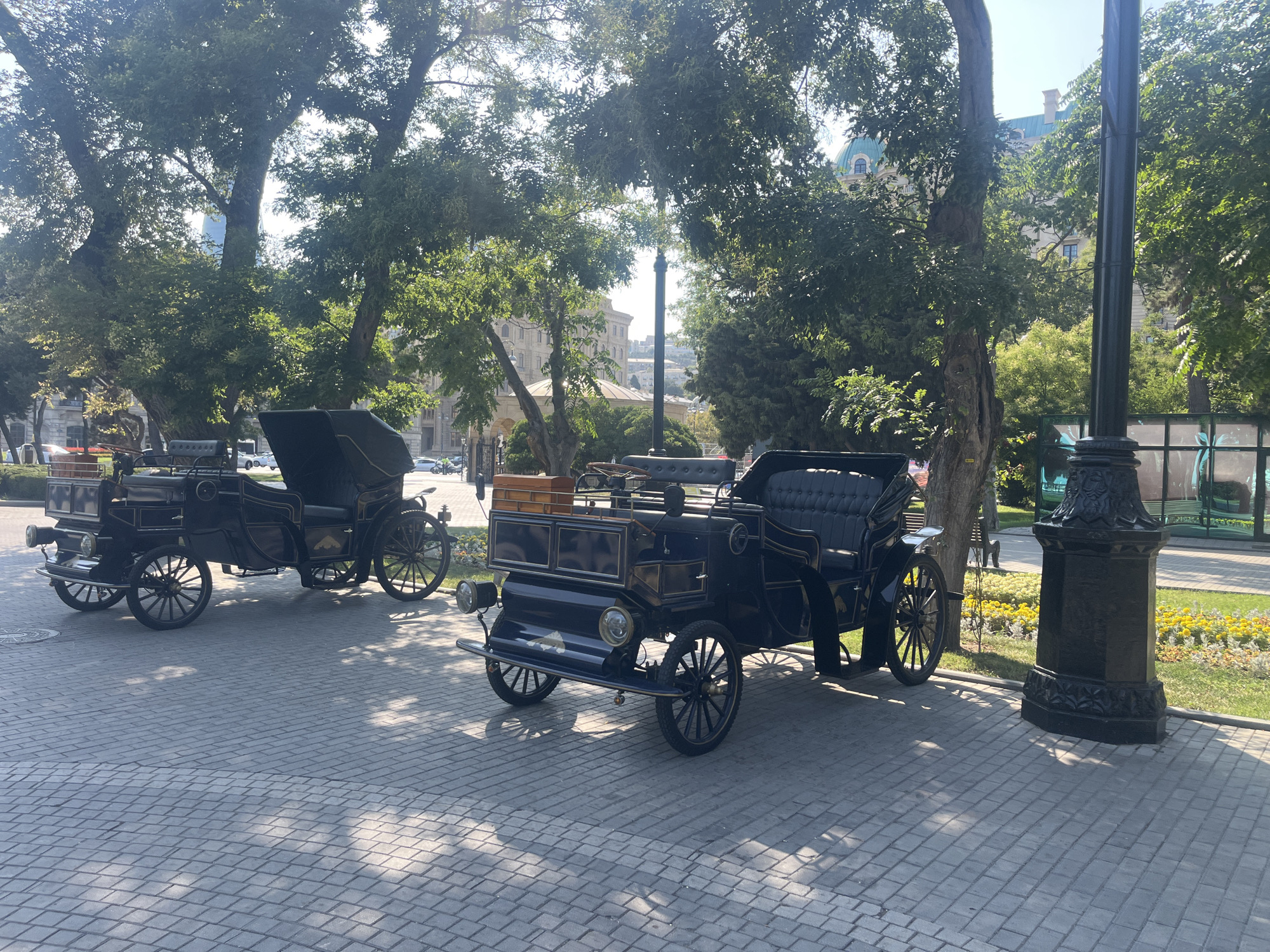 The image captures a sunny day on a paved plaza in Baku, Azerbaijan, with two vintage horse-drawn carriages parked on the cobblestone. The carriages are dark in color, featuring intricate designs and seating for passengers. In the background, mature trees with lush green foliage cast dappled shadows on the ground. Beyond the trees, a tall building with architectural details and a dome can be seen, hinting at the urban environment. The plaza also has a decorative lamppost and a well-maintained garden with yellow flowers. The scene is serene and appears to be a tourist or recreational area.